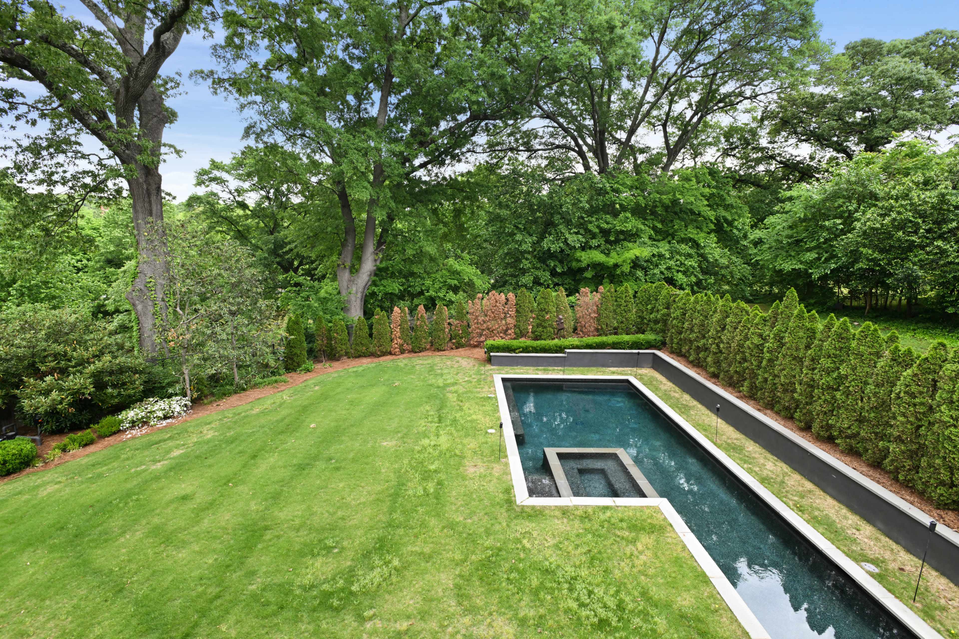 The image shows a landscaped yard featuring a rectangular swimming pool surrounded by a green lawn and tall trees.