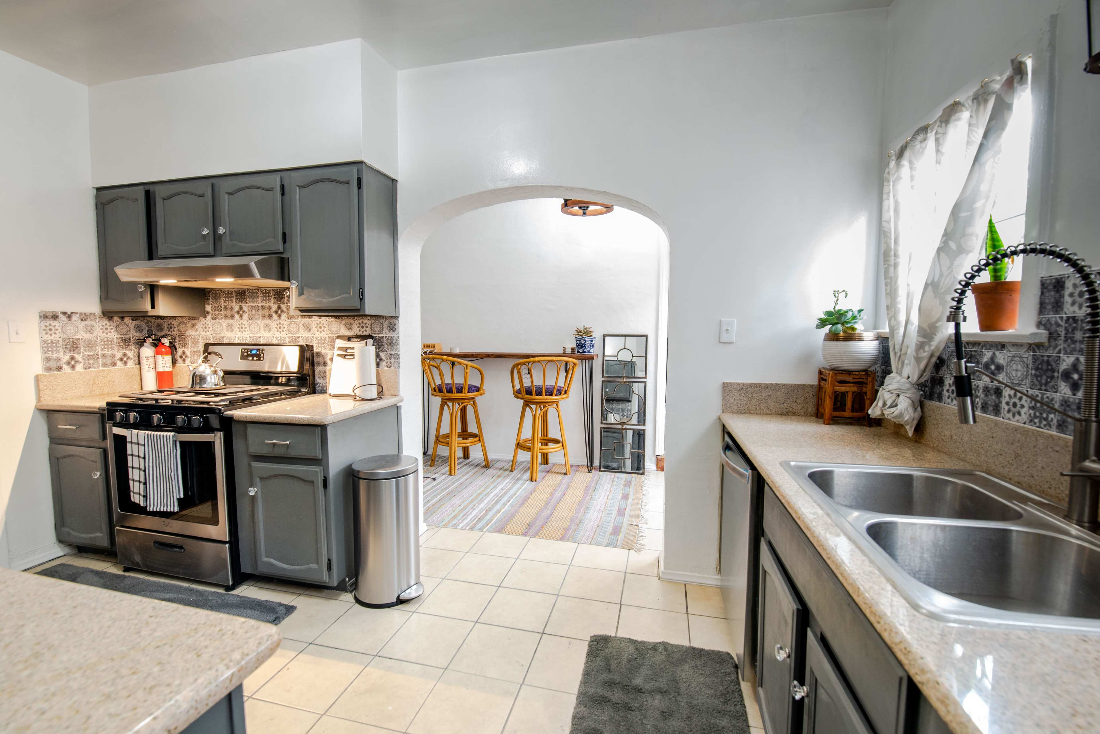 A kitchen features gray cabinets, a gas stove, and a tile floor, with a doorway leading to a dining area with bar stools.