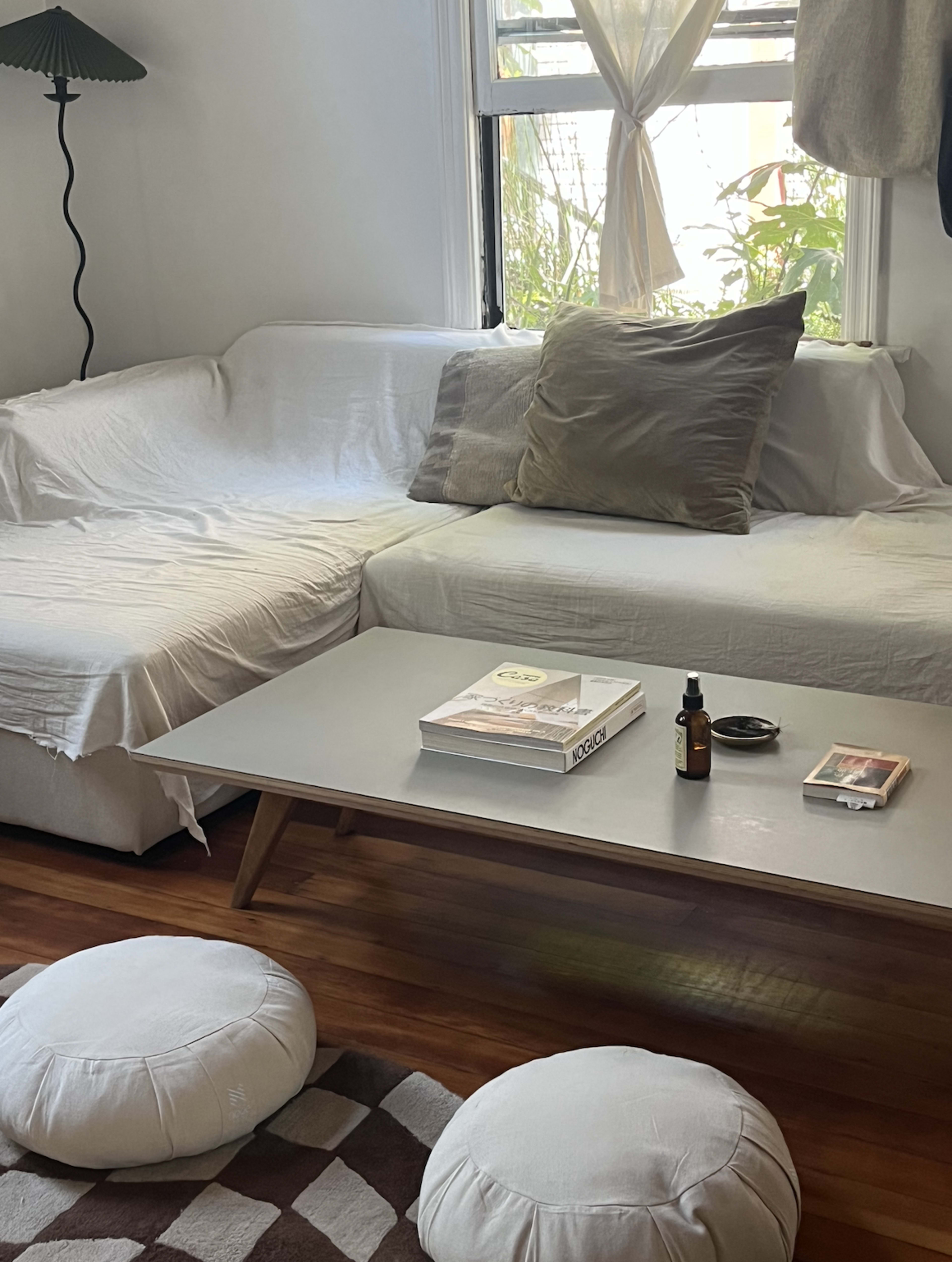 A living room features a white sofa with pillows, a light wooden coffee table displaying a magazine and a small bottle, and two round cushions on a patterned rug.