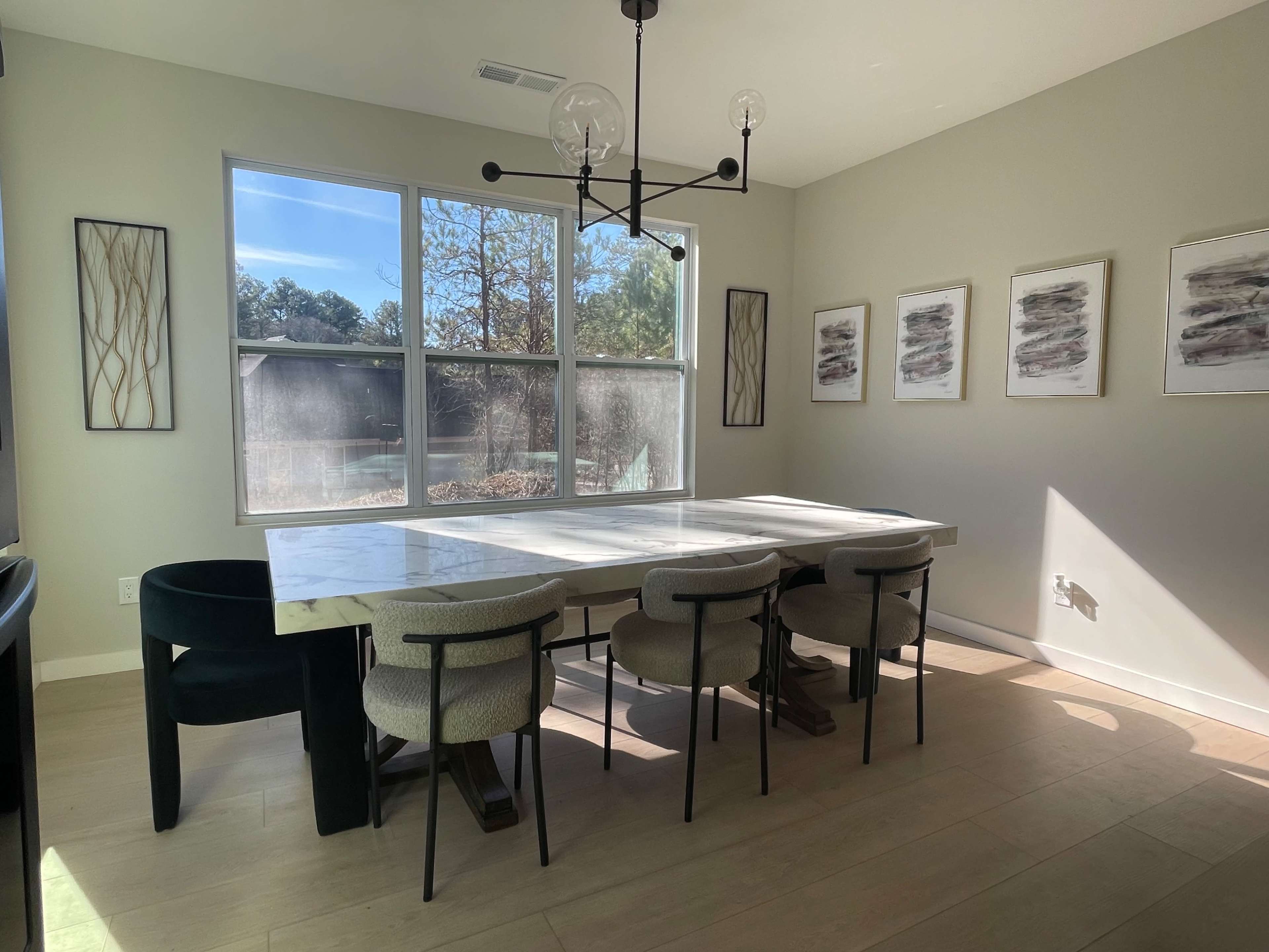 A dining room features a large marble table surrounded by chairs and a modern hanging light fixture above, with large windows letting in natural light.