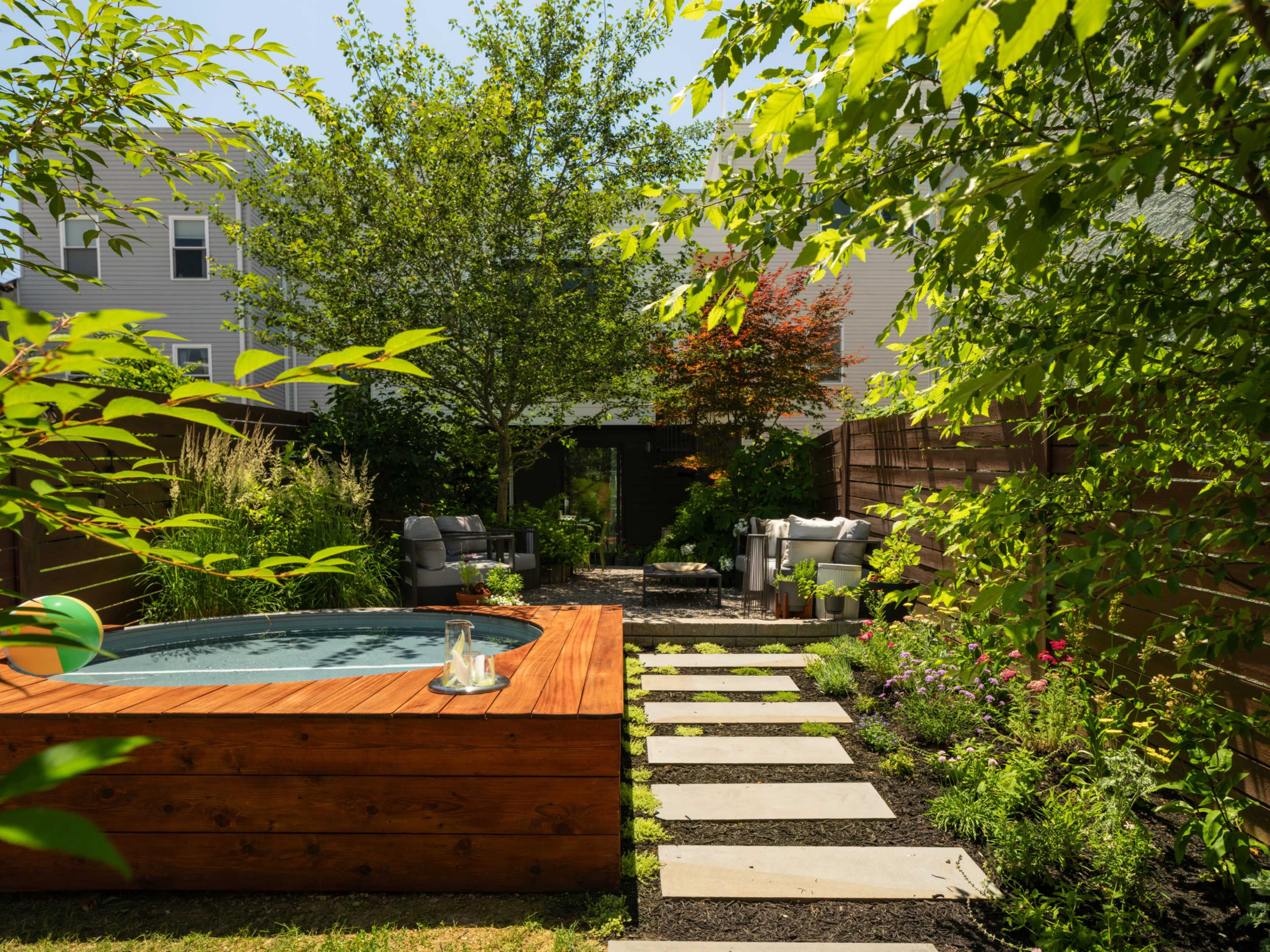 A wooden hot tub is surrounded by landscaped greenery and stone pathways in a backyard garden.