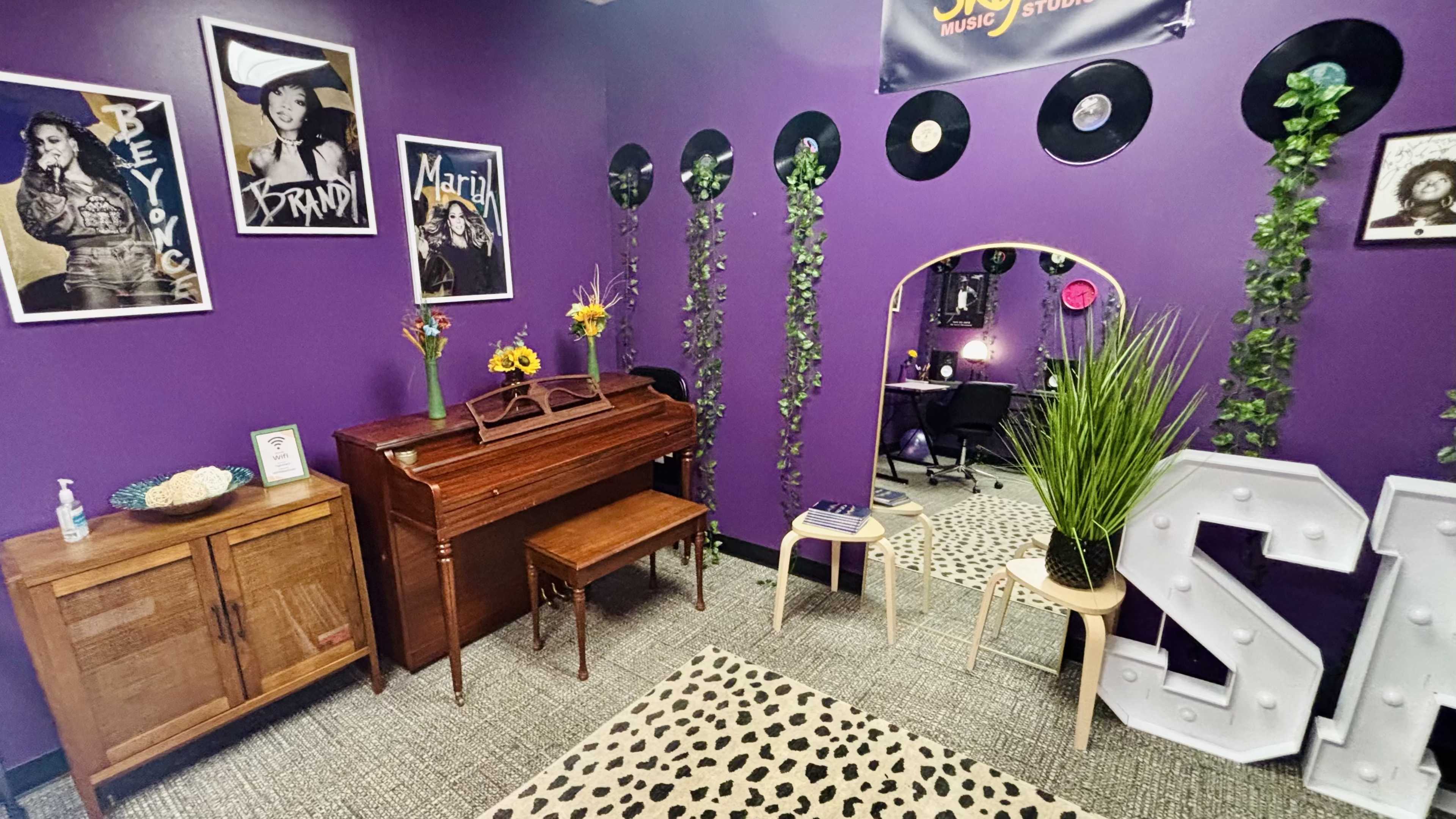 The image shows a vibrant purple room decorated with music-themed posters, vinyl records on the walls, and a vintage piano accompanied by plants and flowers.