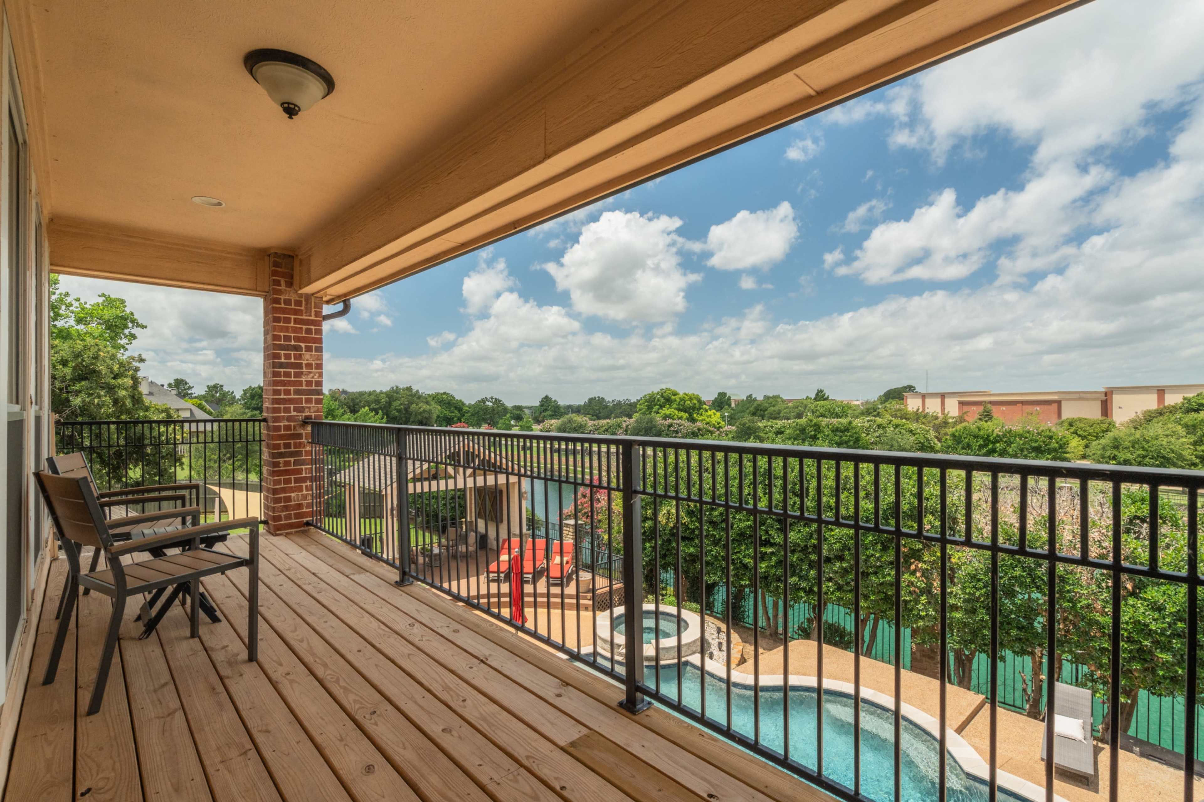 A spacious balcony overlooks a pool area surrounded by greenery under a partly cloudy sky.