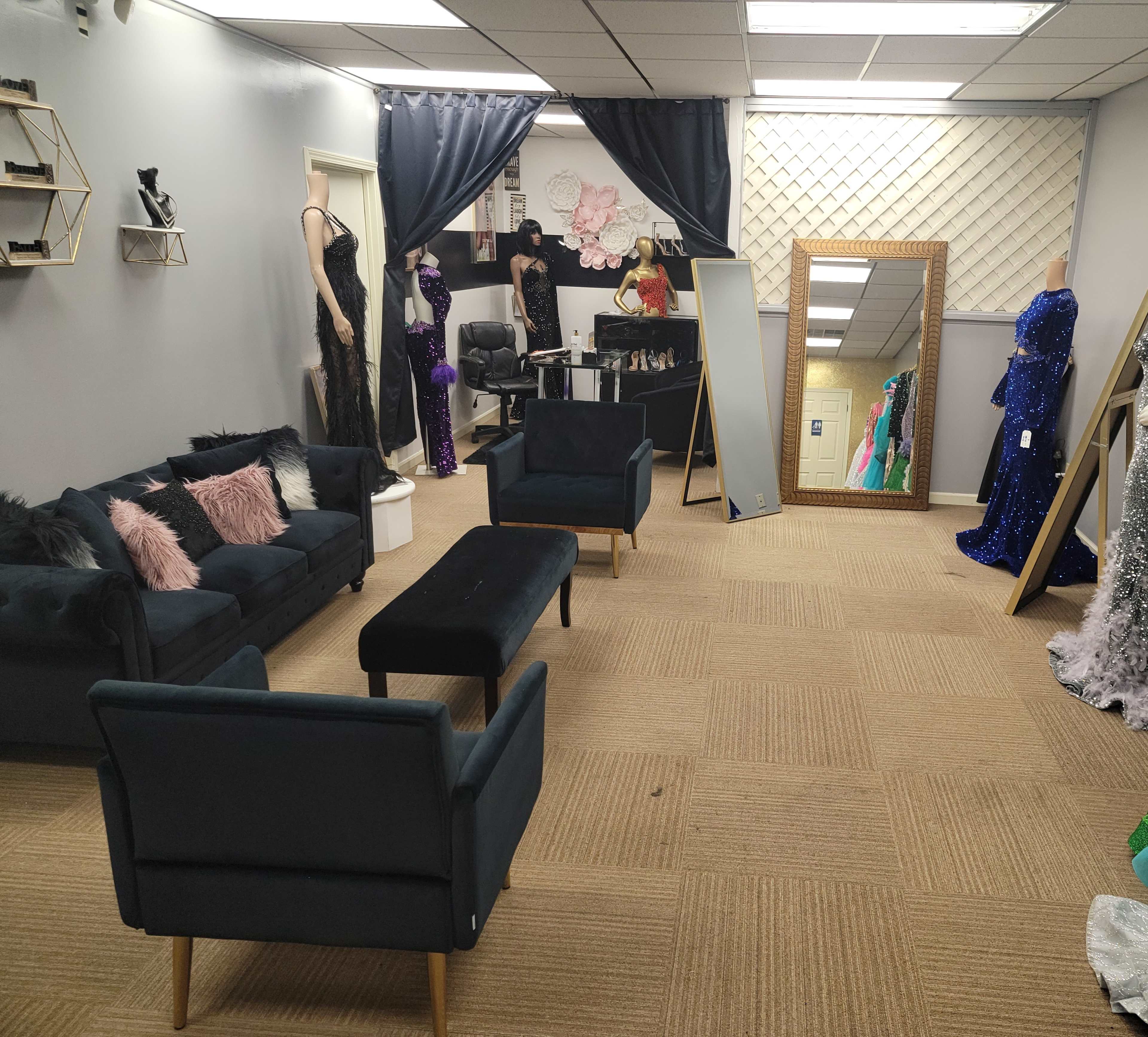 A boutique interior with dark green sofas, a mirror, and various formal dresses displayed on racks and mannequins.
