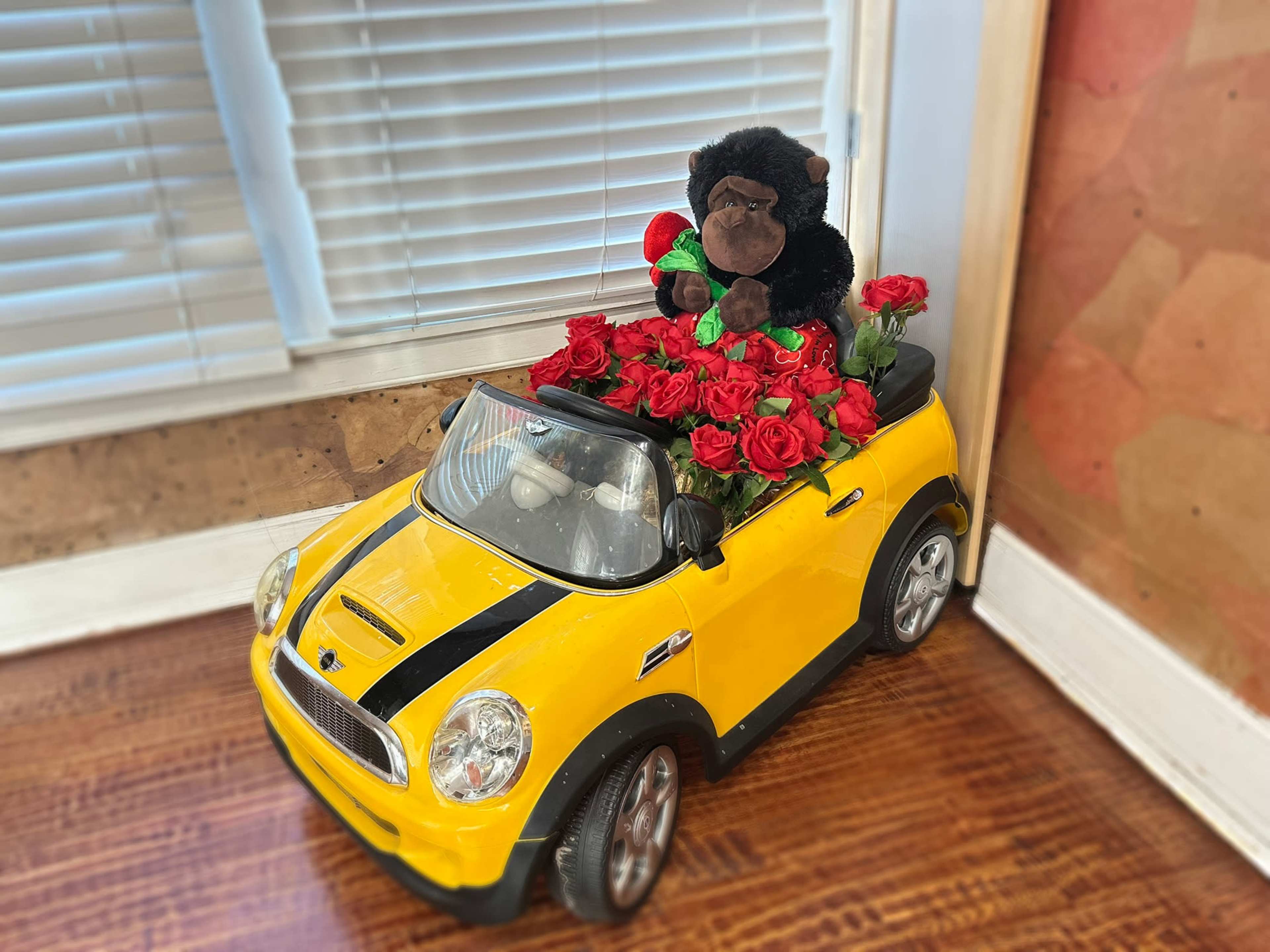 A toy yellow car filled with red roses and featuring a black plush gorilla in the driver's seat is positioned near a window.