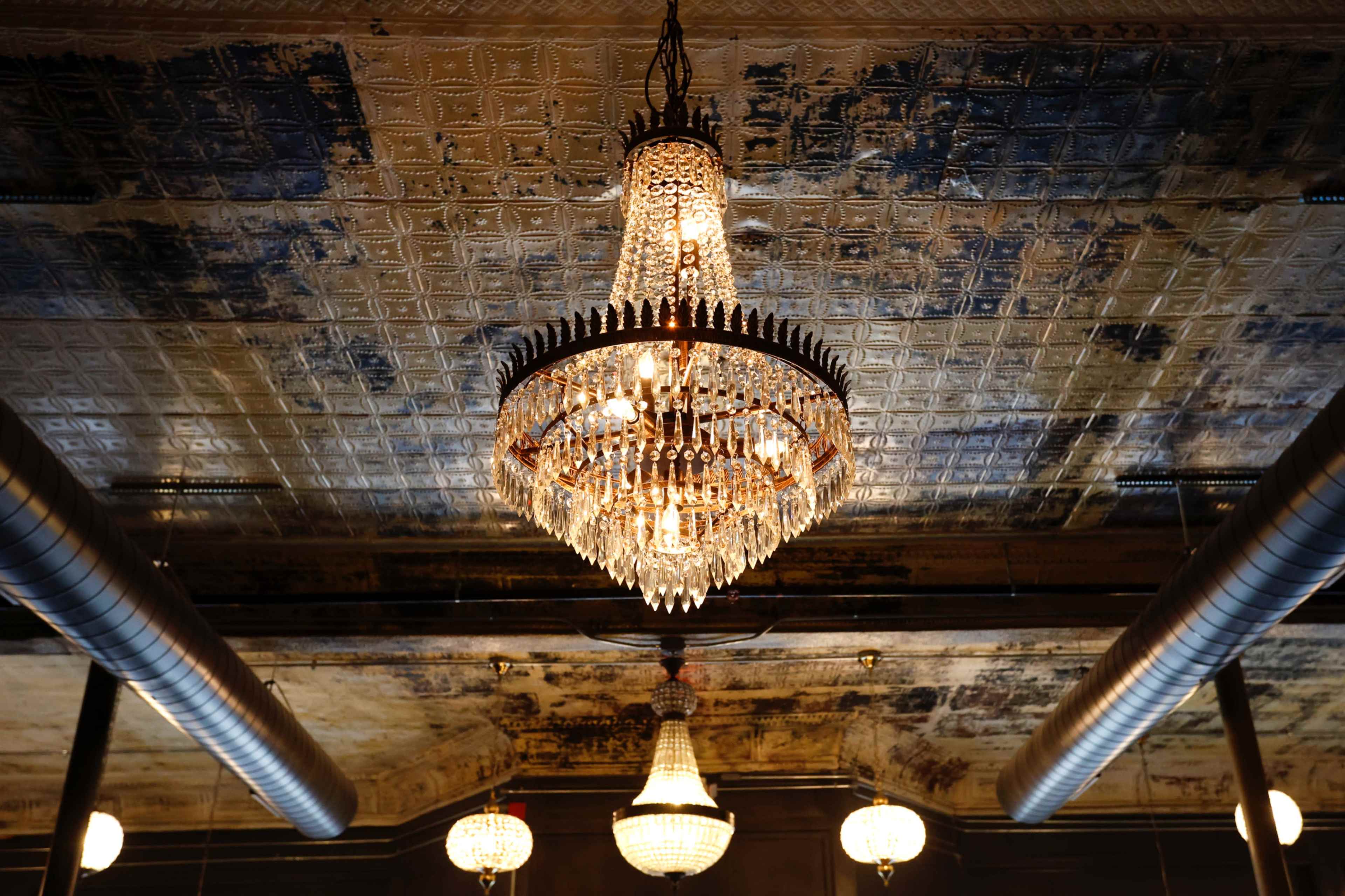 The image shows a chandelier hanging from an ornate, patterned ceiling with visible textures and lighting fixtures around it.