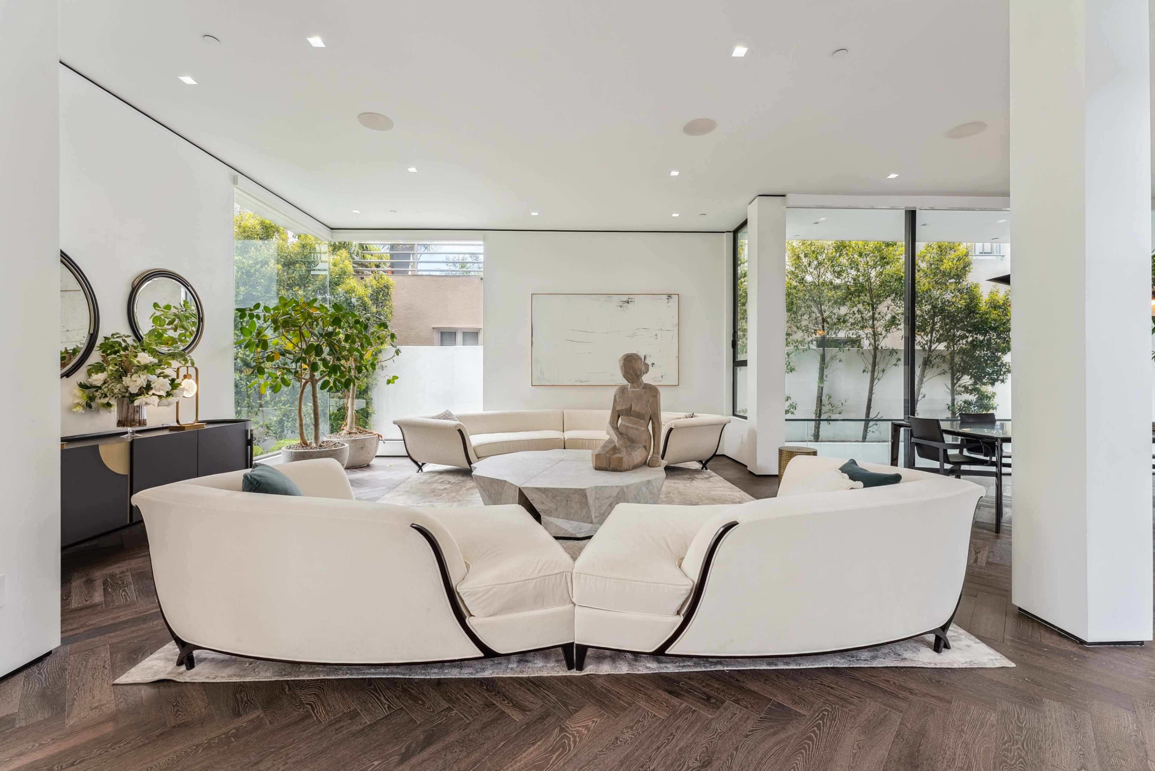 The image shows a modern living room with two curved white sofas facing a central sculpture and surrounded by large windows that provide natural light and views of greenery outside.