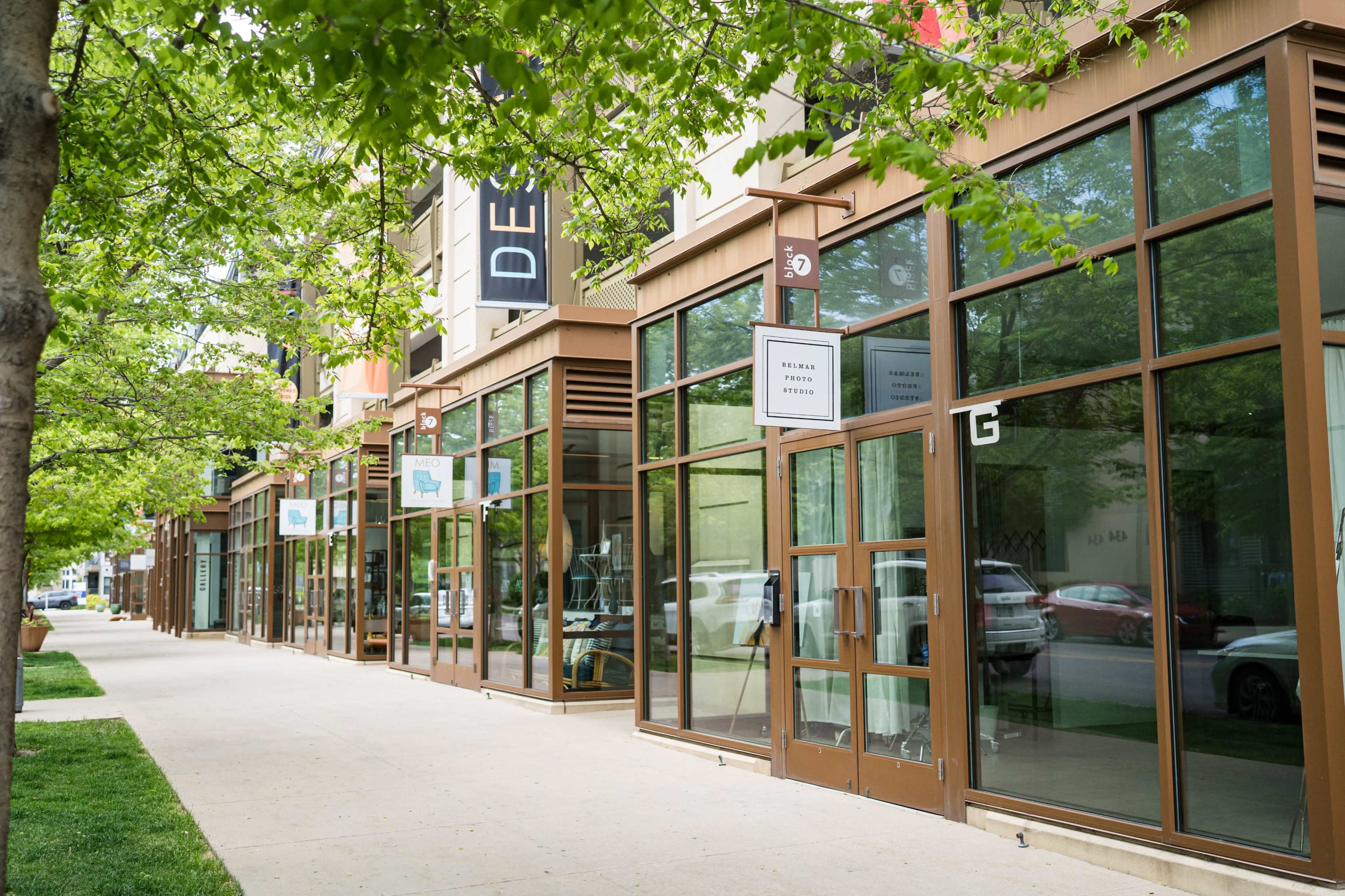 A row of storefronts with large windows and greenery along the sidewalk.