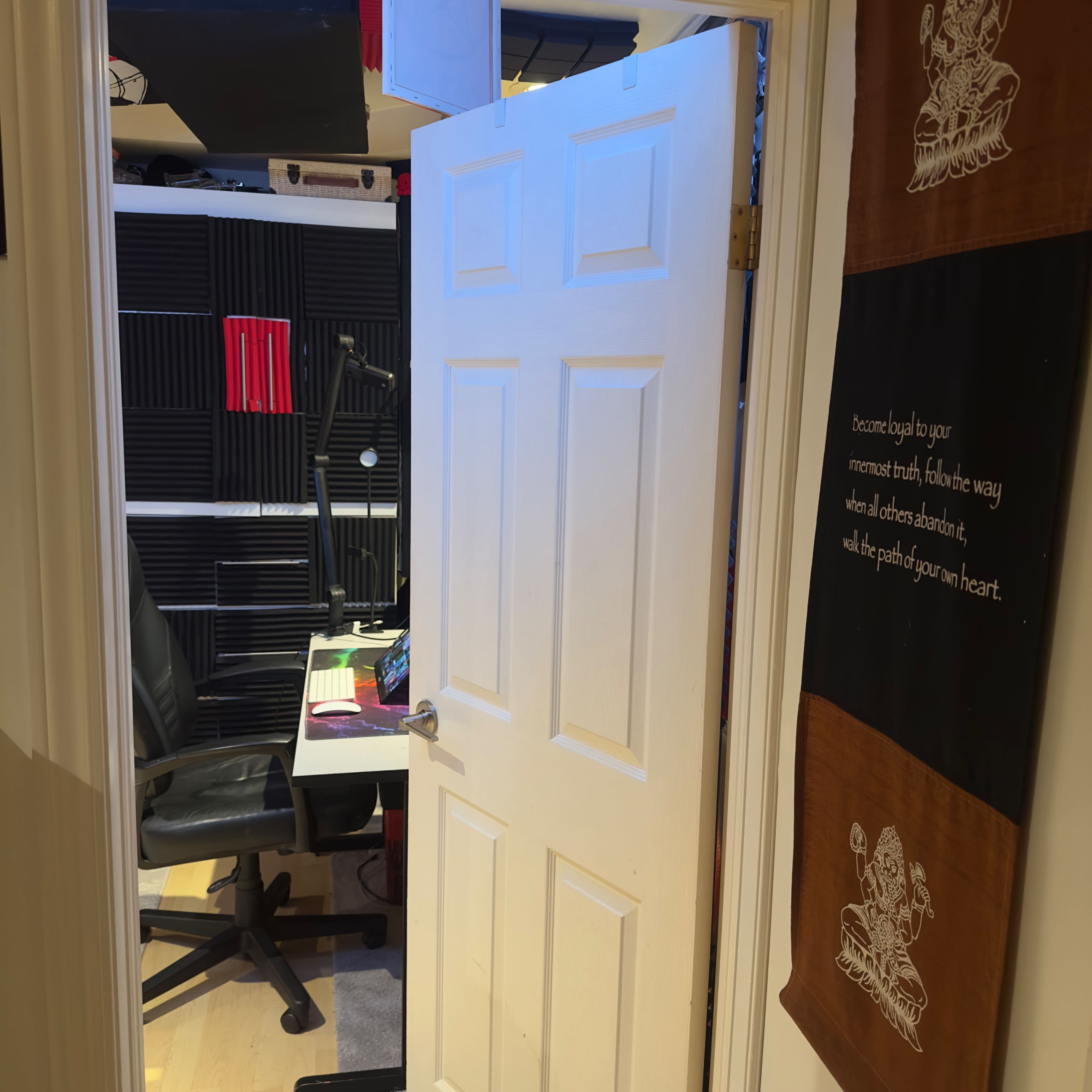The image shows an open doorway leading into a room with a desk, chair, and soundproofing panels on the walls.