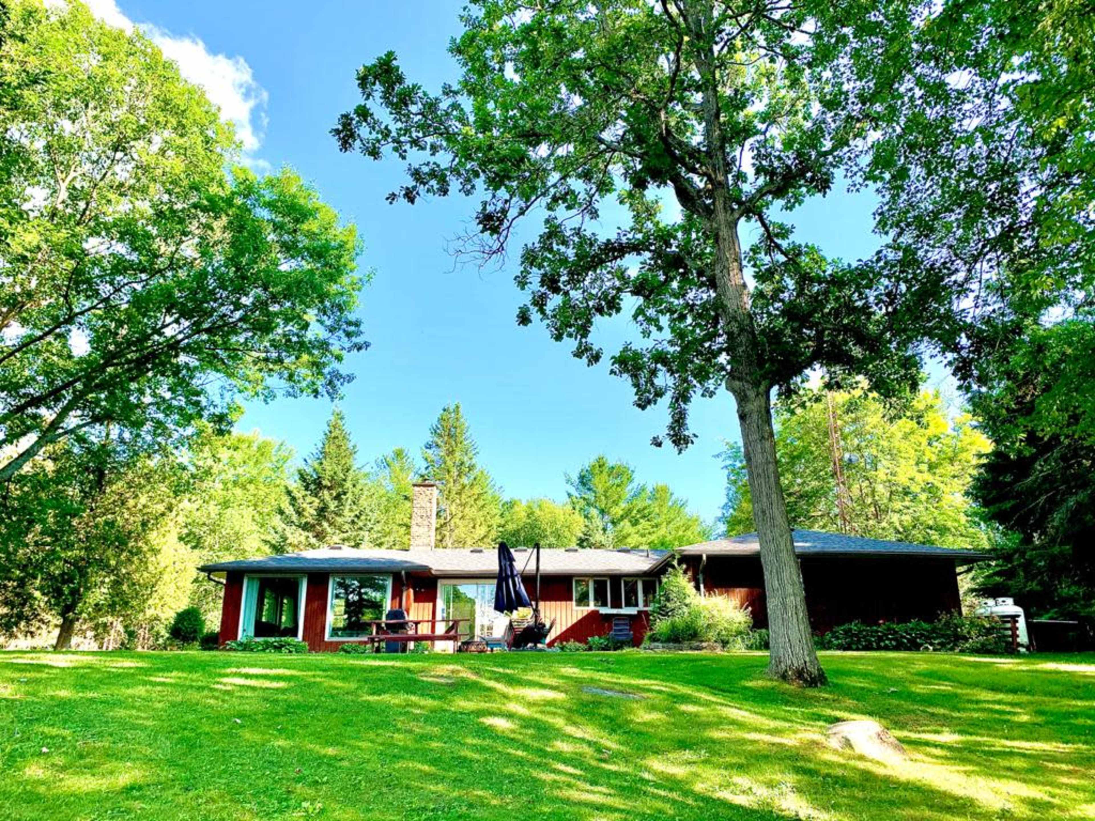 A single-story red house with large windows sits on a green lawn surrounded by trees under a clear blue sky.