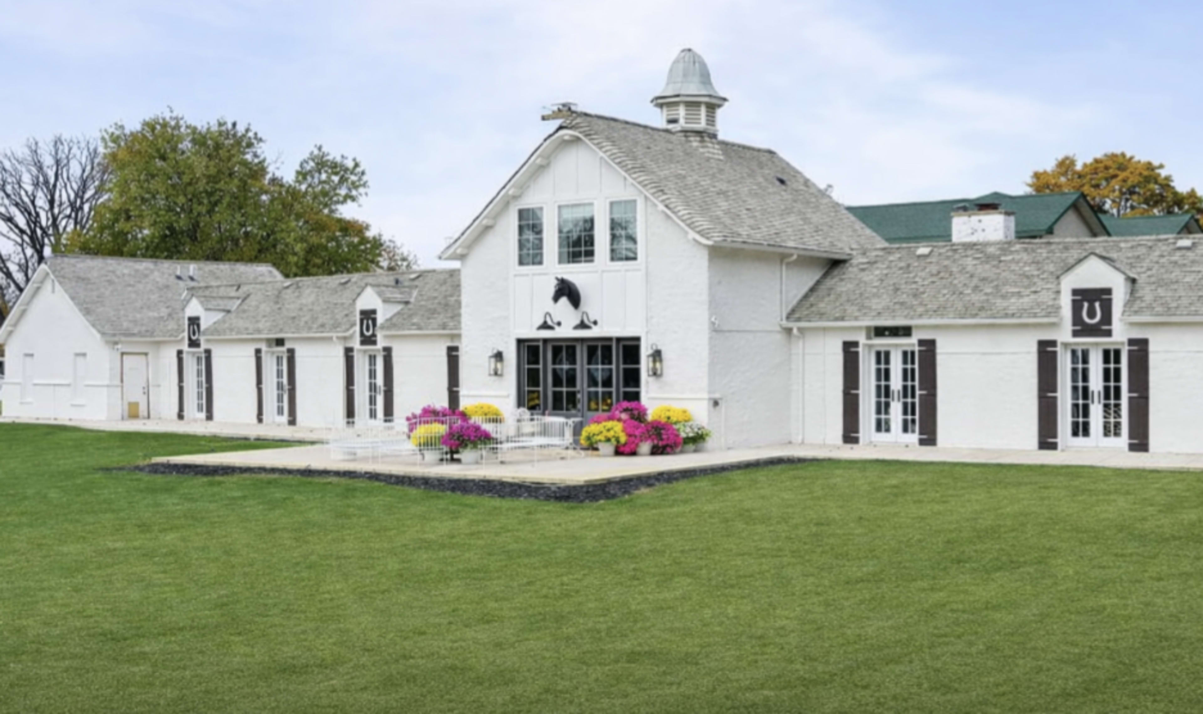 The image shows a spacious white building with a grey roof surrounded by well-maintained green grass and colorful flower arrangements in front.