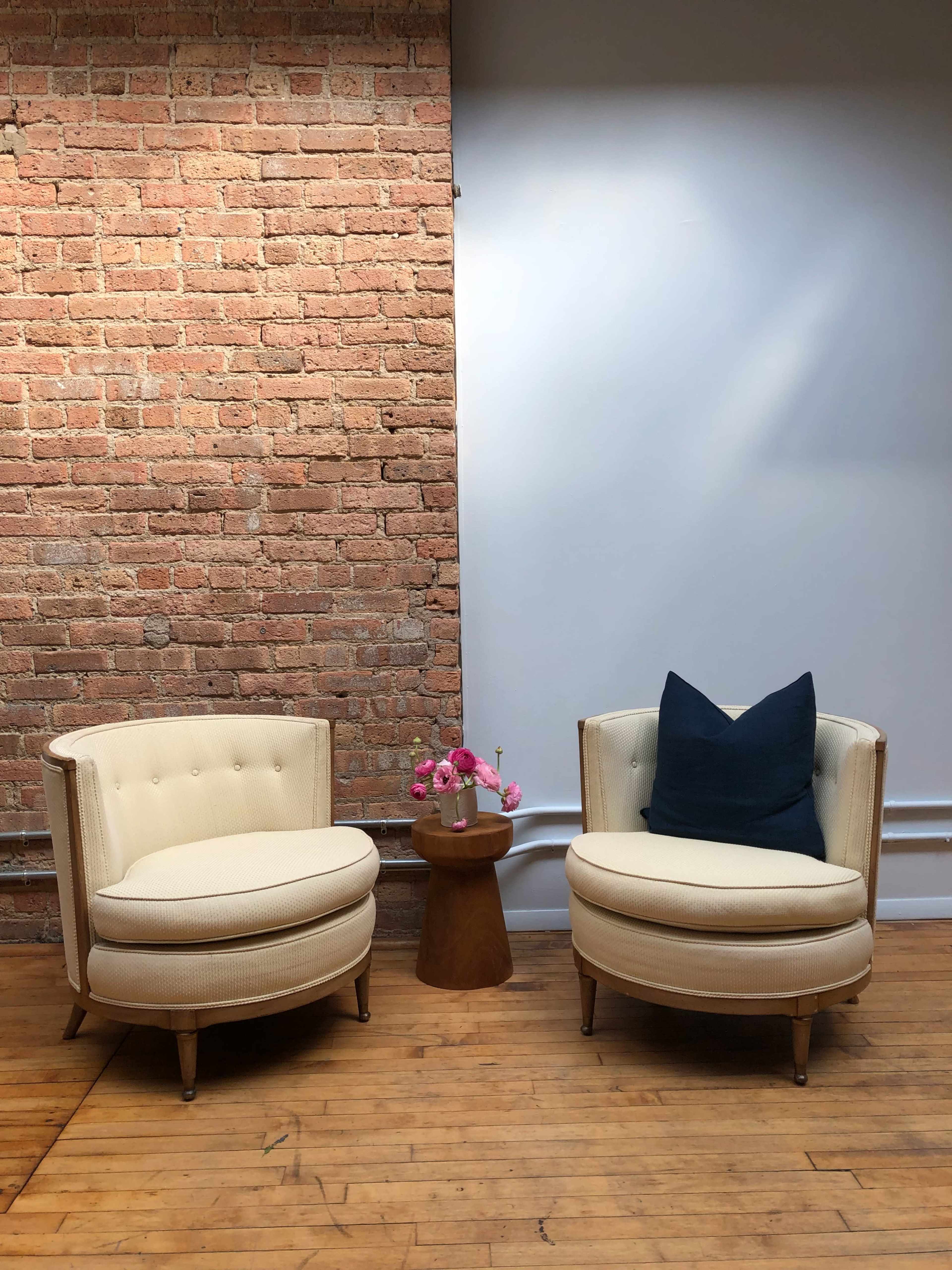 Two cream-colored chairs with round bases are positioned opposite each other, flanking a small wooden table adorned with a vase of pink flowers against a brick wall.