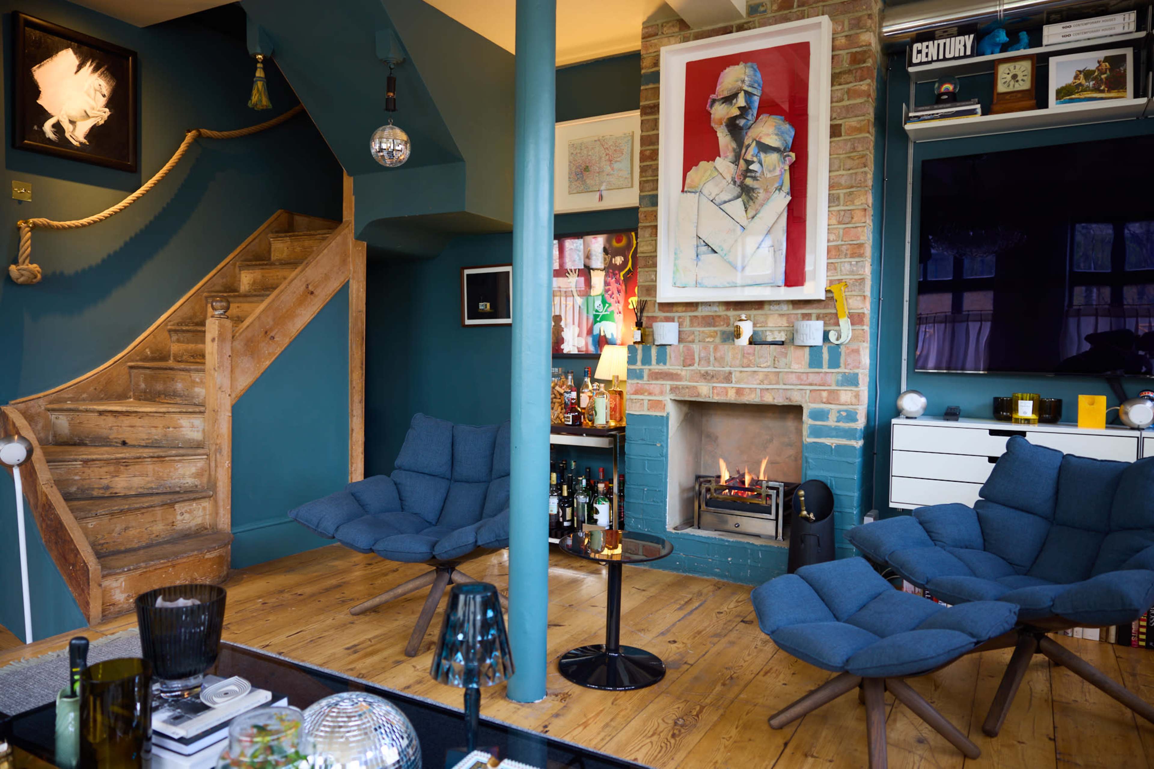 A cozy living room with a fireplace, wooden flooring, blue chairs, and a staircase leading to an upper level.