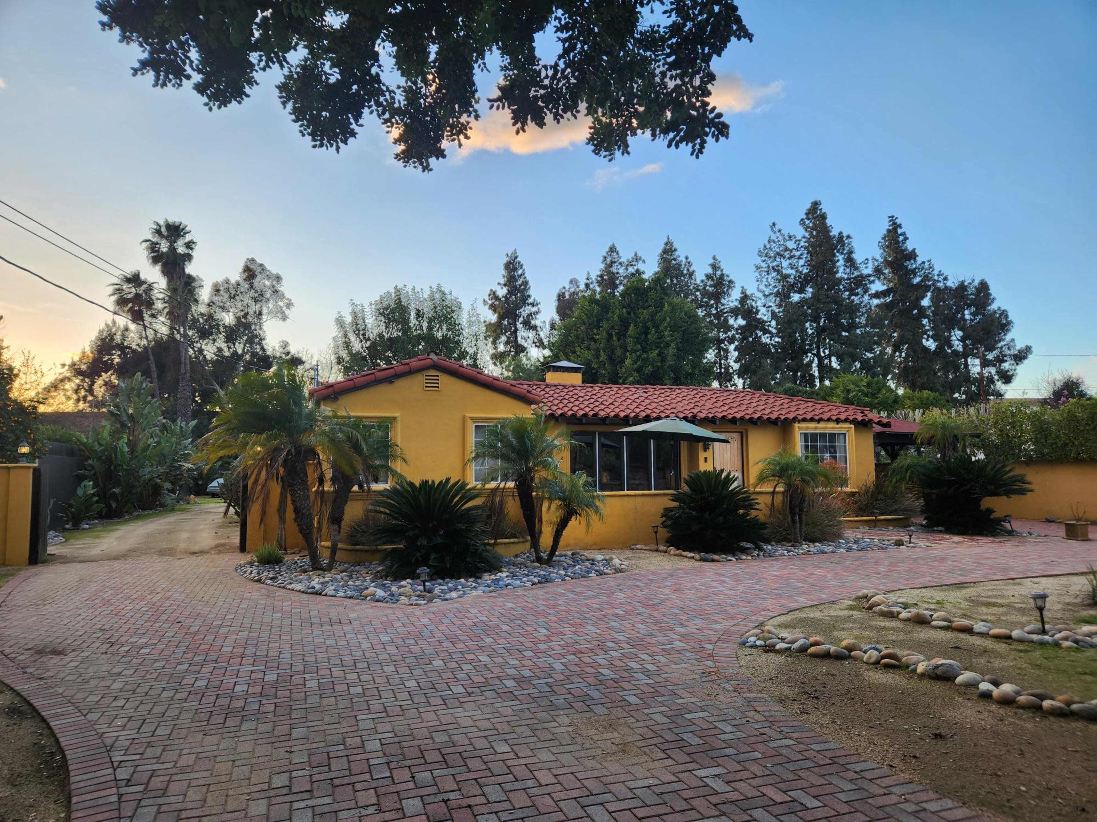 A yellow, single-story house with a red tiled roof is surrounded by palm trees and a circular brick driveway.