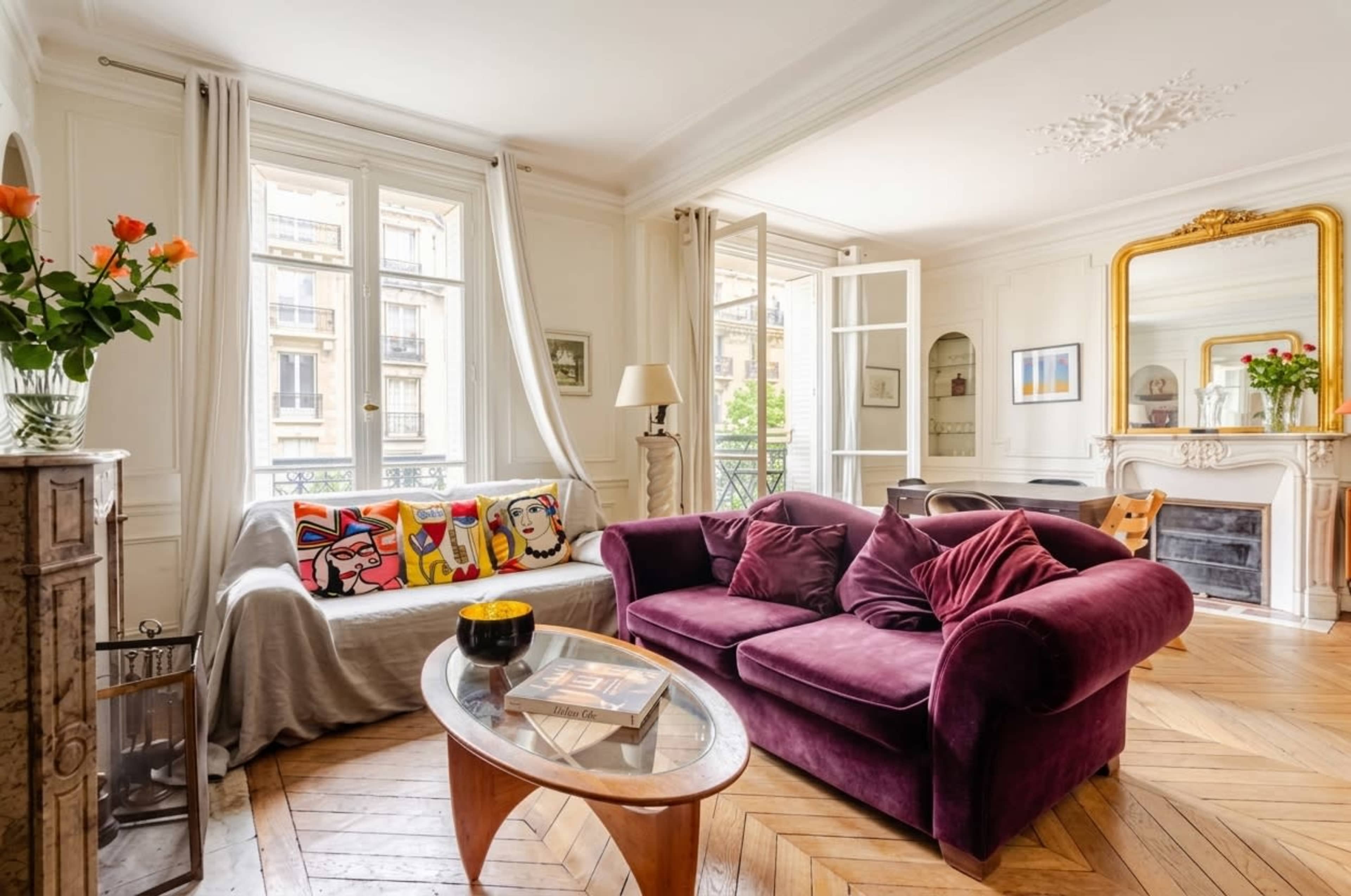 A living room features a purple sofa, a glass coffee table, and a decorative fireplace, with large windows that open to a balcony.