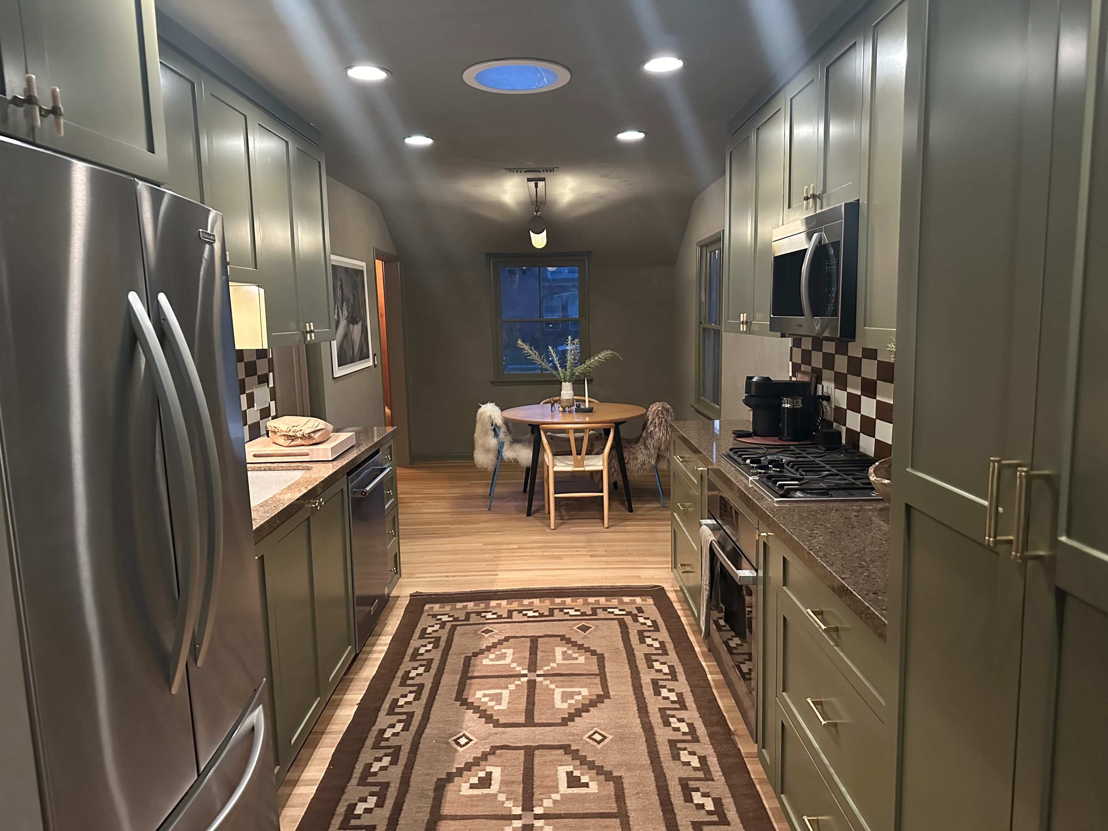 The image shows a modern kitchen with green cabinetry, stainless steel appliances, a patterned rug, and a dining table by a window.