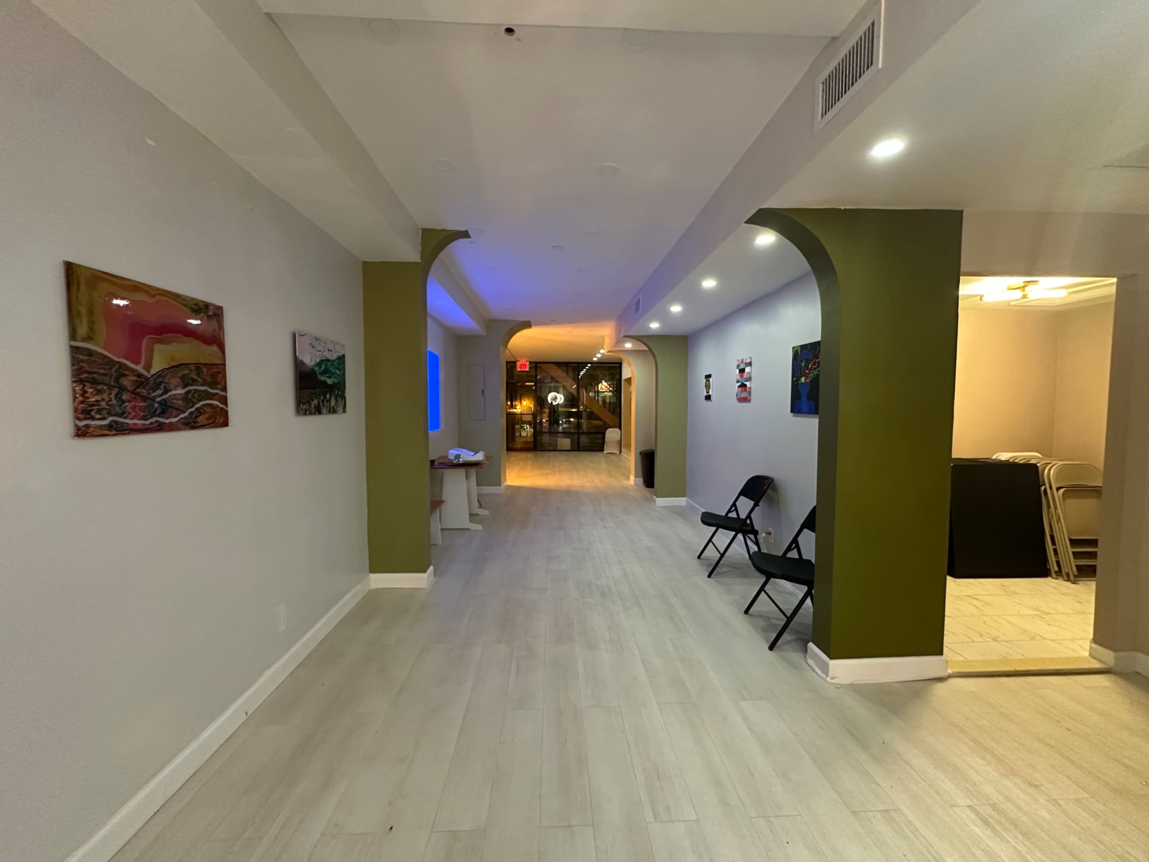 The image shows a wide, well-lit hallway in a modern interior space with artworks displayed on the walls and chairs set up along one side.