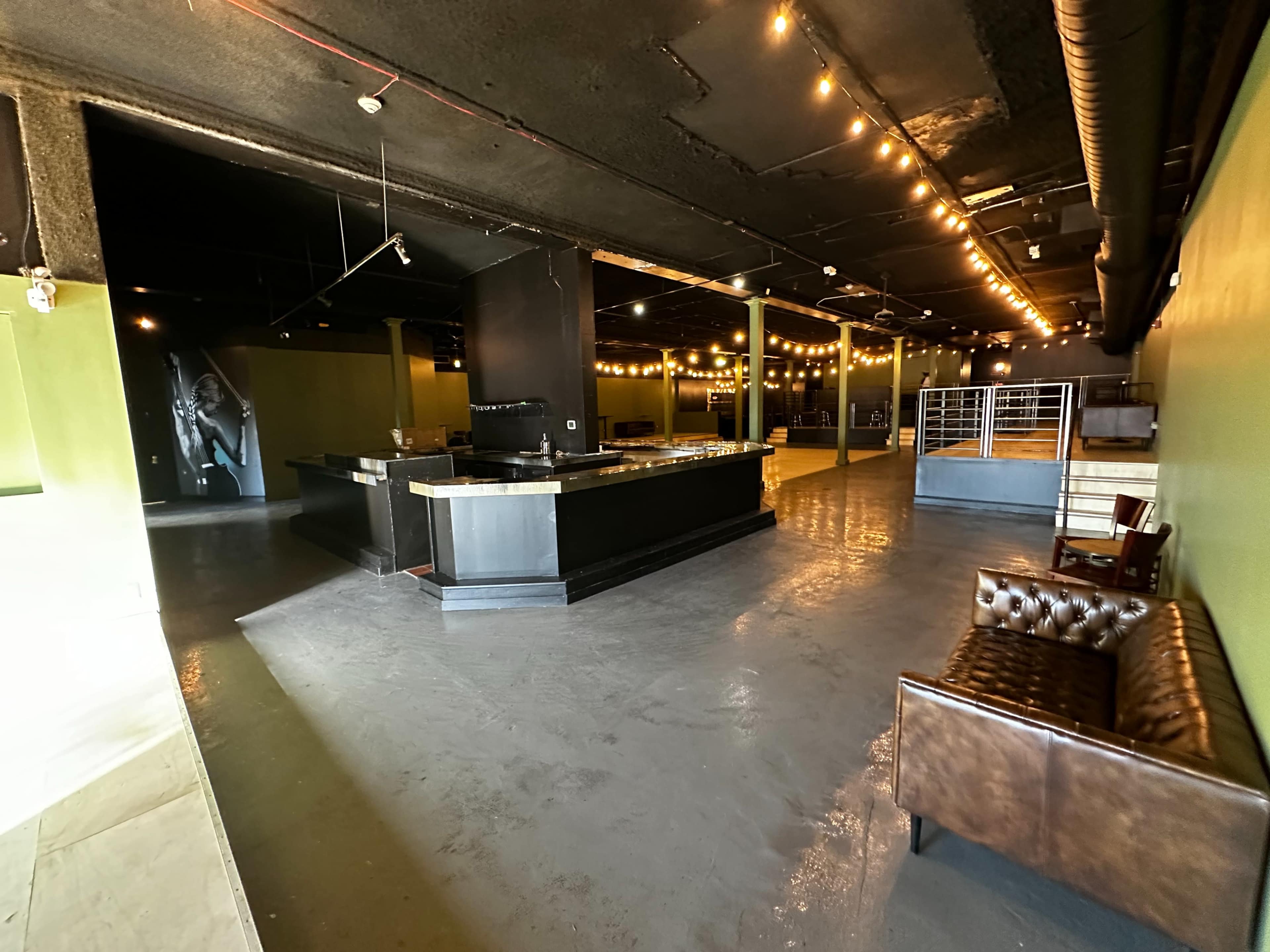 A spacious, dimly lit interior of a commercial venue featuring a bar area, exposed ceilings with hanging lights, and an empty seating area.