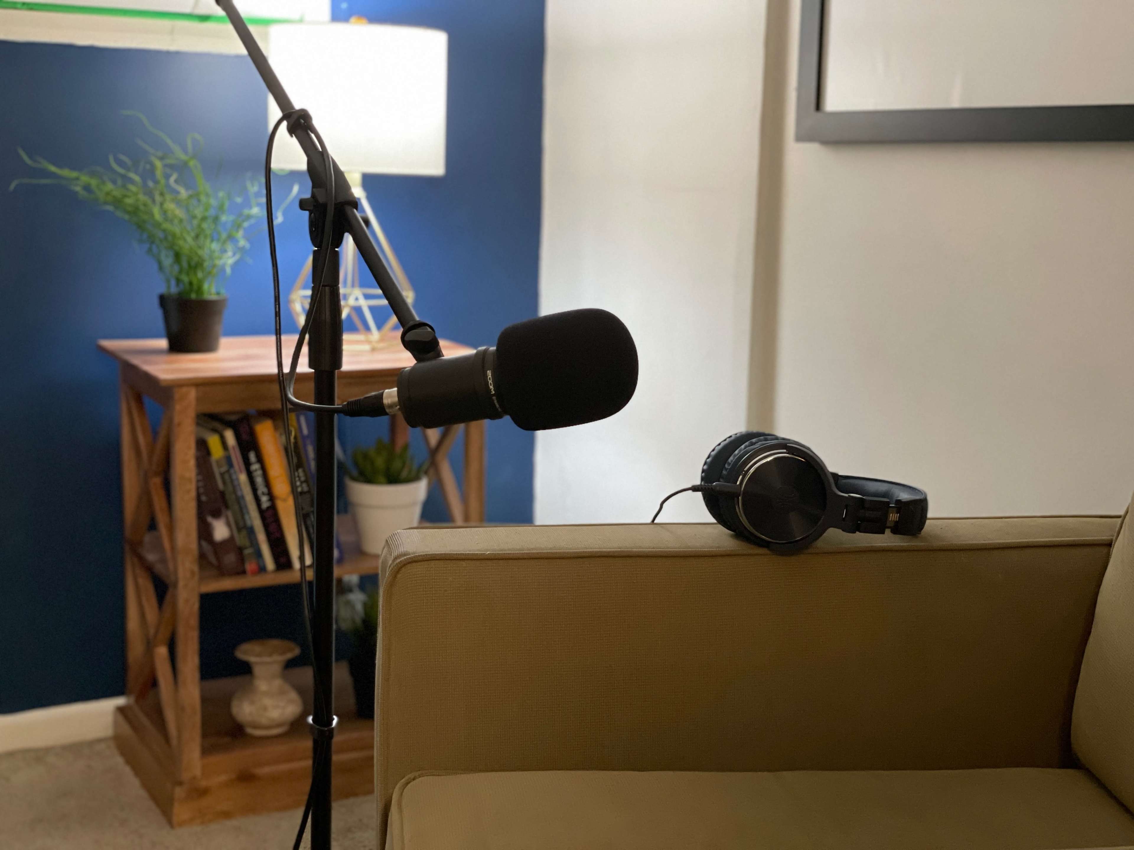 A microphone on a stand is positioned near a couch with headphones resting on the arm, beside a bookshelf and a potted plant.