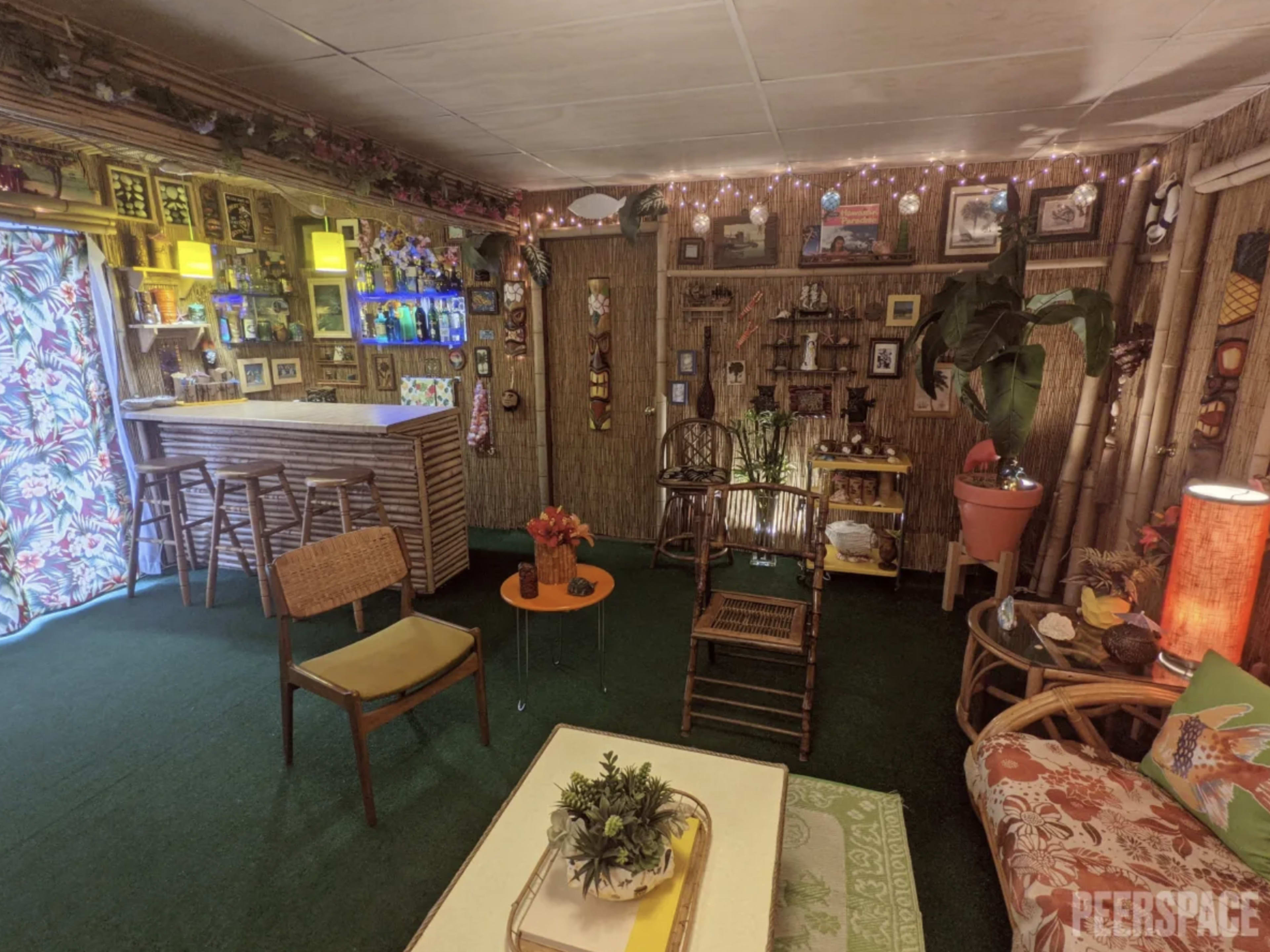 The image shows a tiki-themed lounge area with bamboo decor, a bar, and various plants, illuminated by soft lighting and adorned with tropical artwork.