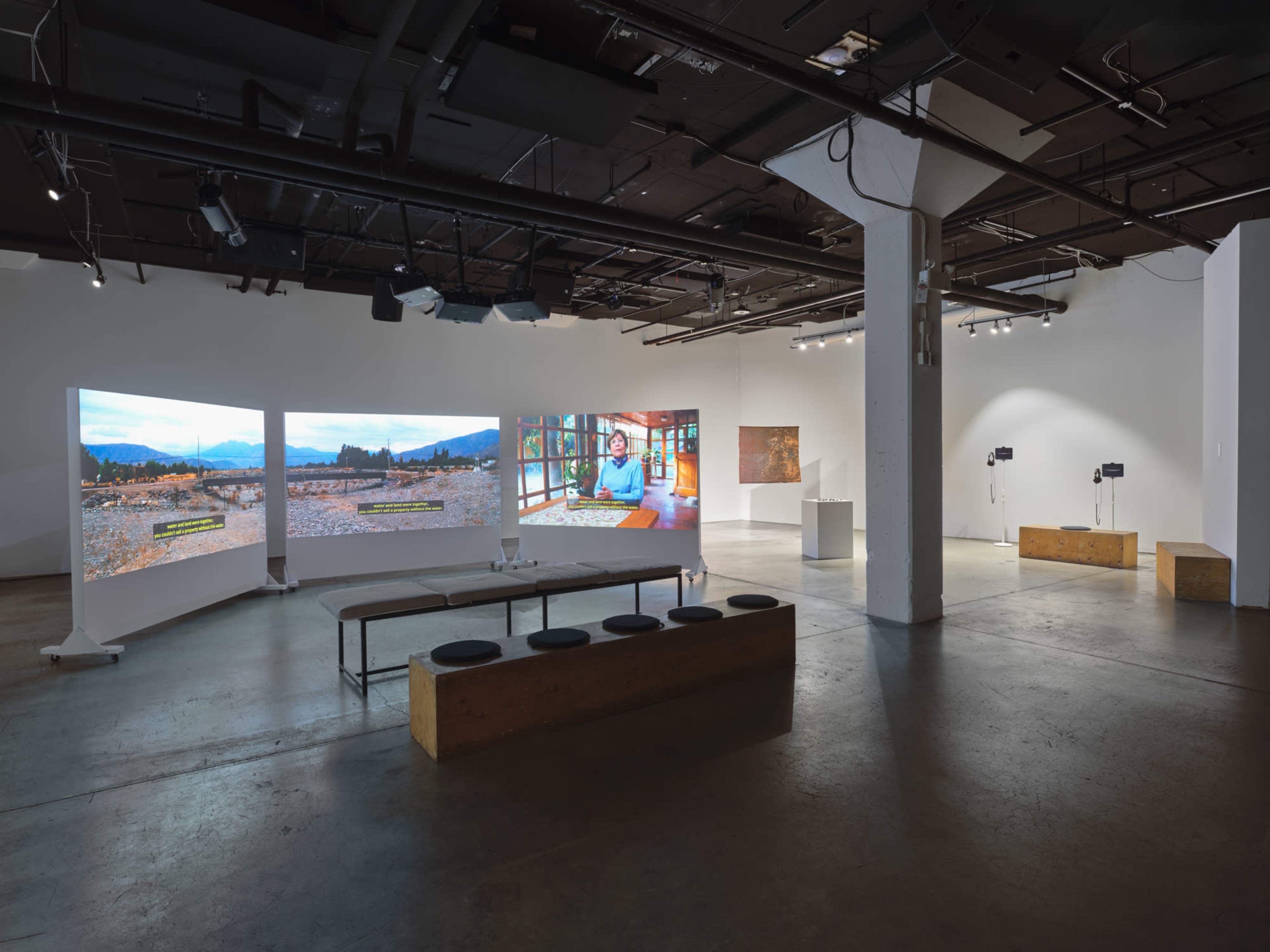 The image shows an art gallery with large screens displaying videos, a central bench, and minimal decor in a spacious room.