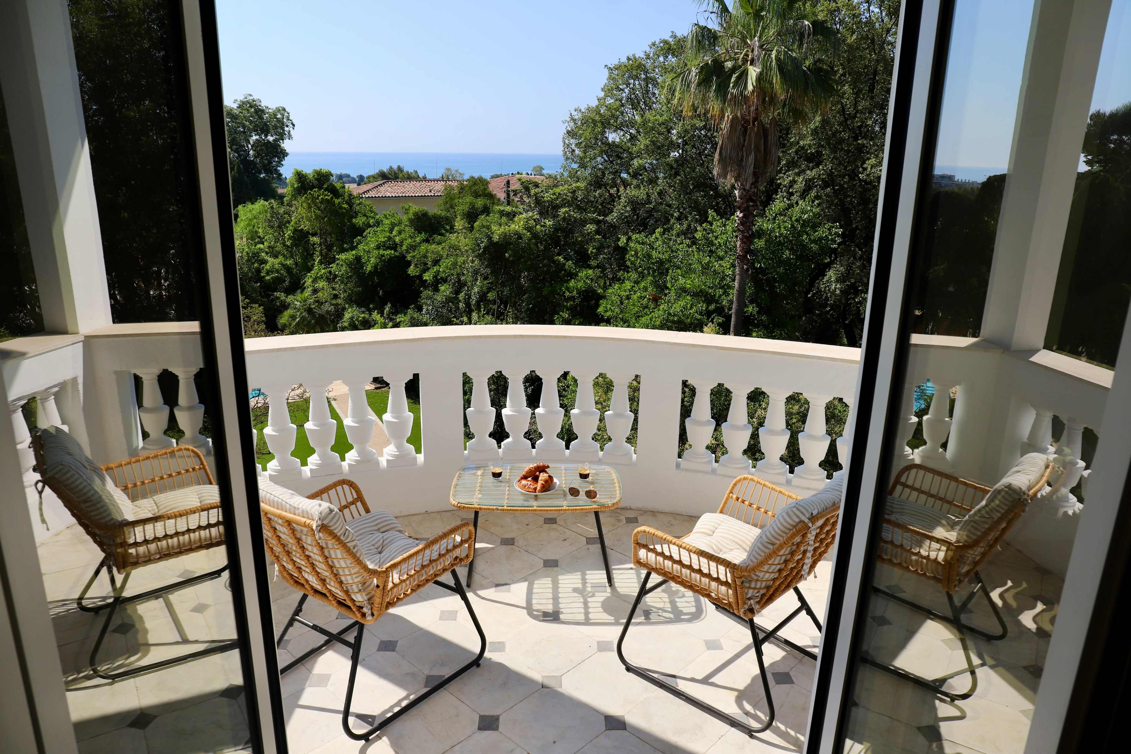 A balcony features two wicker chairs and a glass table set against a backdrop of lush greenery and ocean views.
