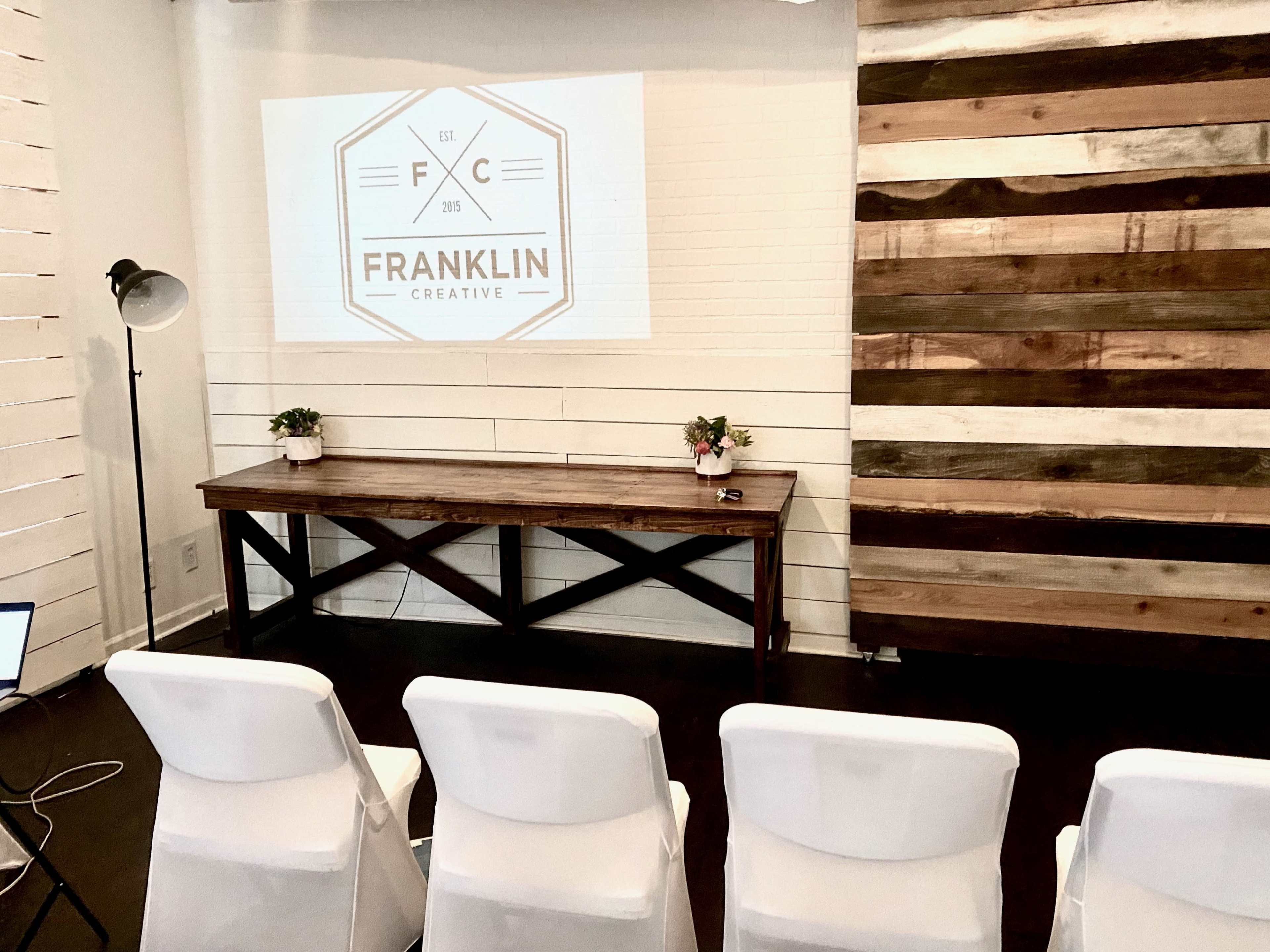A presentation setup with a wooden table, chairs, and a projector displaying the "FRANKLIN CREATIVE" logo on a white wall.