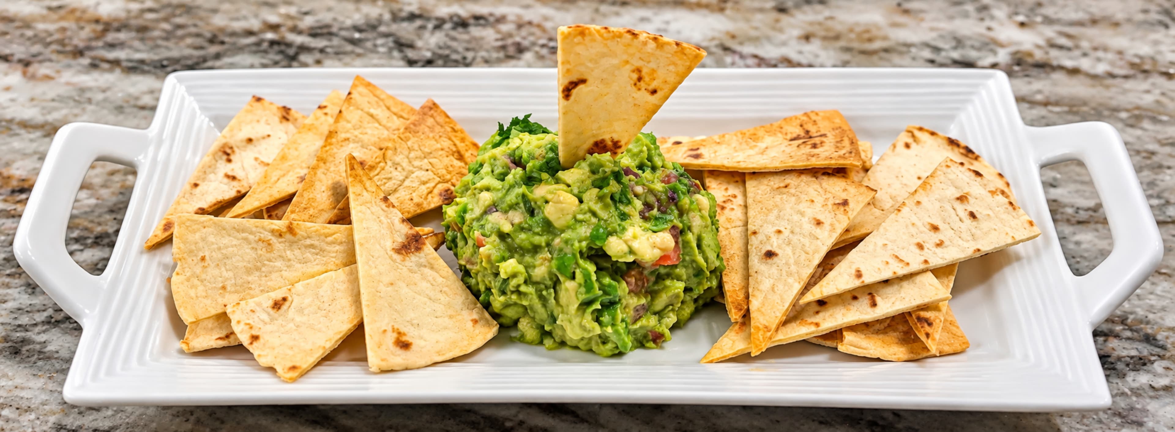 A platter of tortilla chips surrounds a central serving of guacamole on a white rectangular dish.