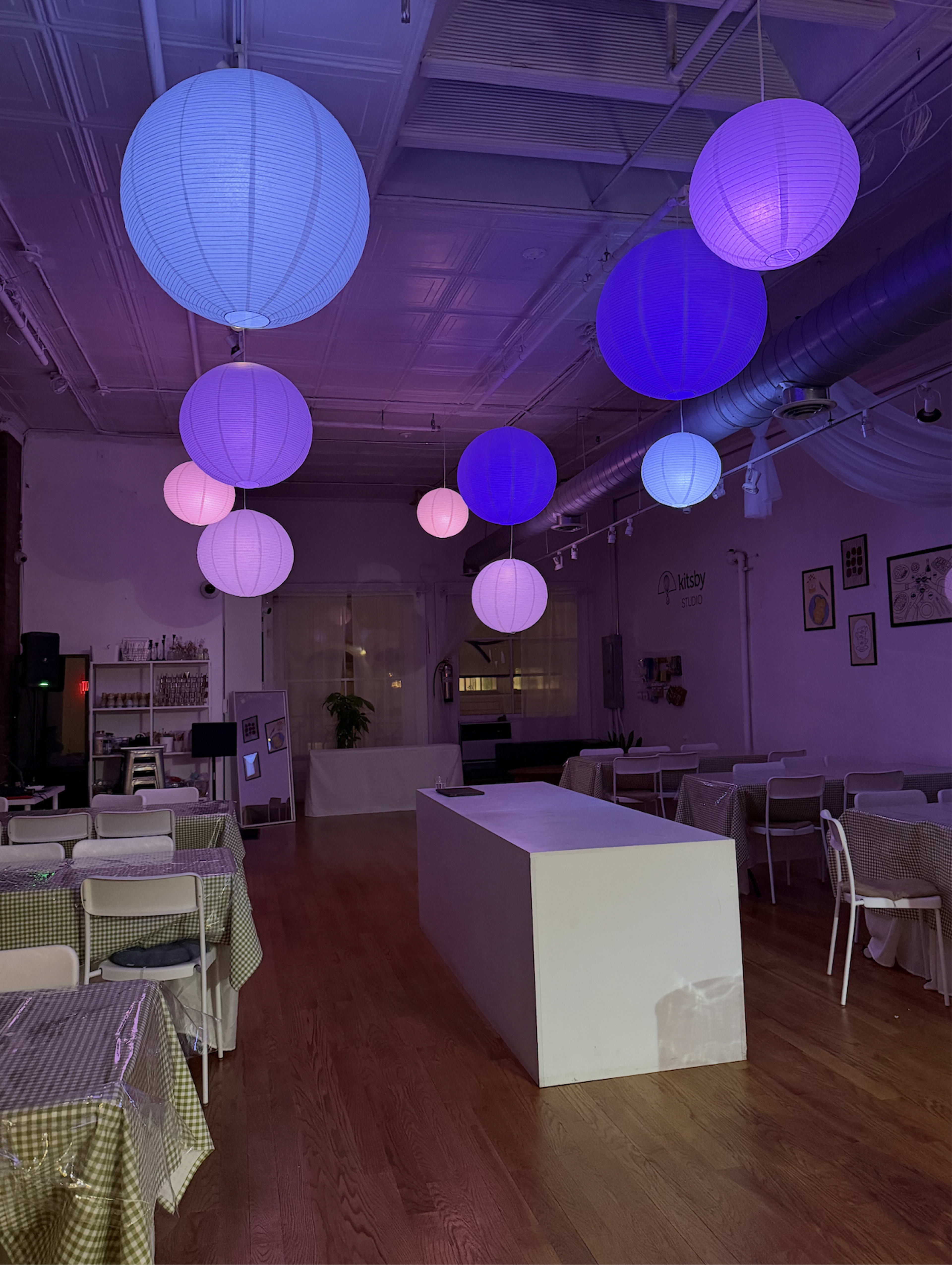 The image shows a decorative indoor venue with several illuminated paper lanterns hanging from the ceiling, positioned above tables covered in checkered tablecloths.