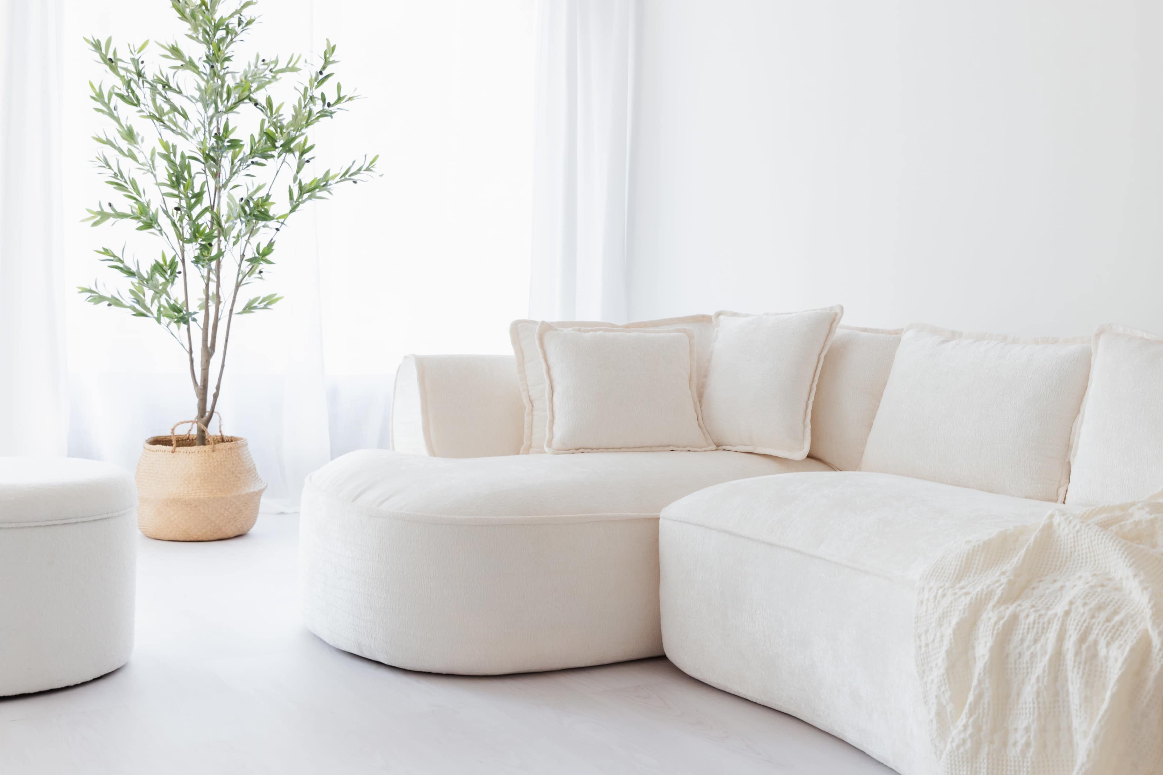 The image depicts a light-colored, plush sectional sofa with two accent pillows, accompanied by a potted plant in a woven basket, all set against a bright, minimalistic interior with white walls and large windows.