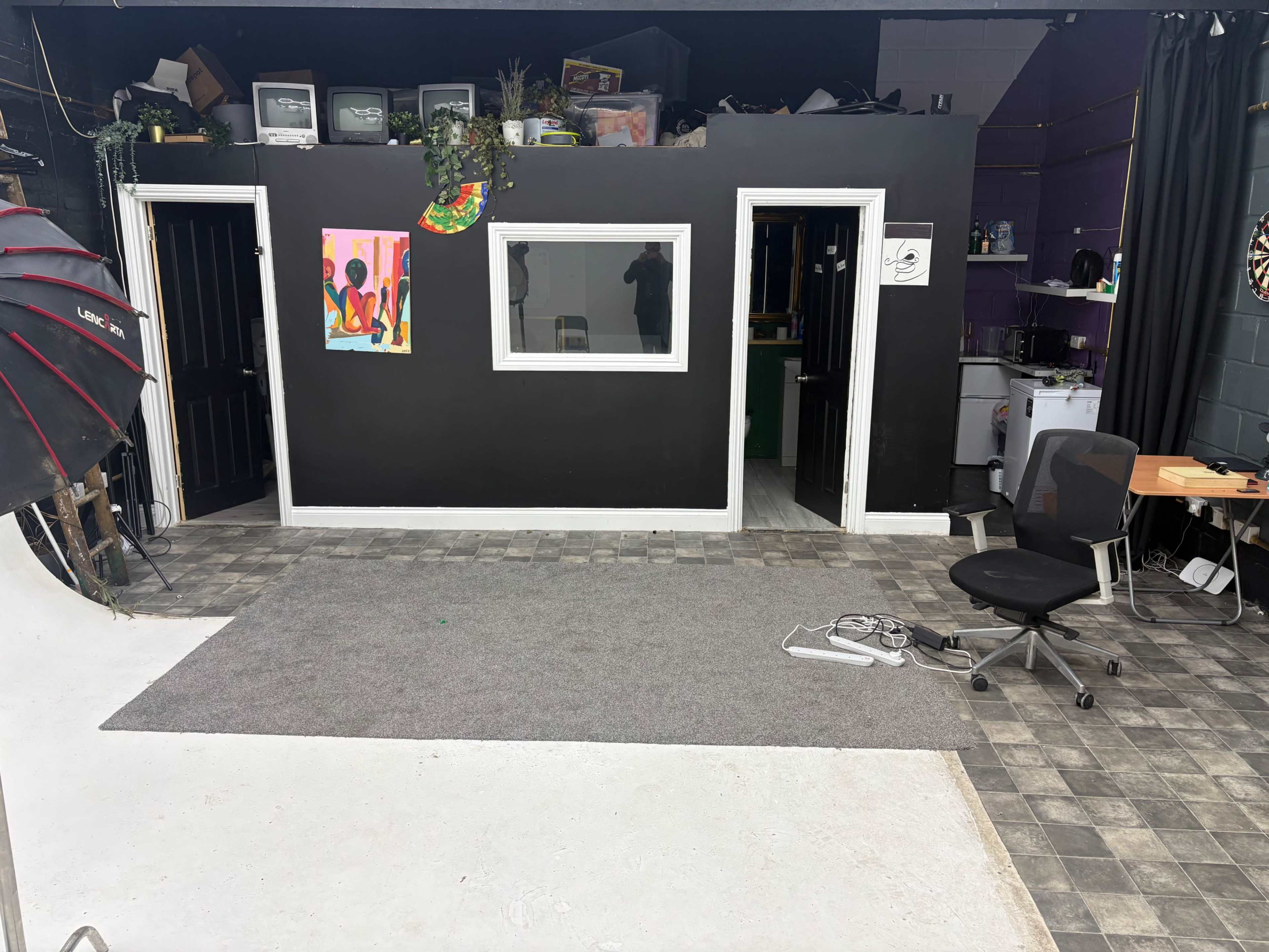 The image shows an indoor studio space with black walls, two doorways, a colorful artwork on the wall, and a gray carpet covering part of the floor.