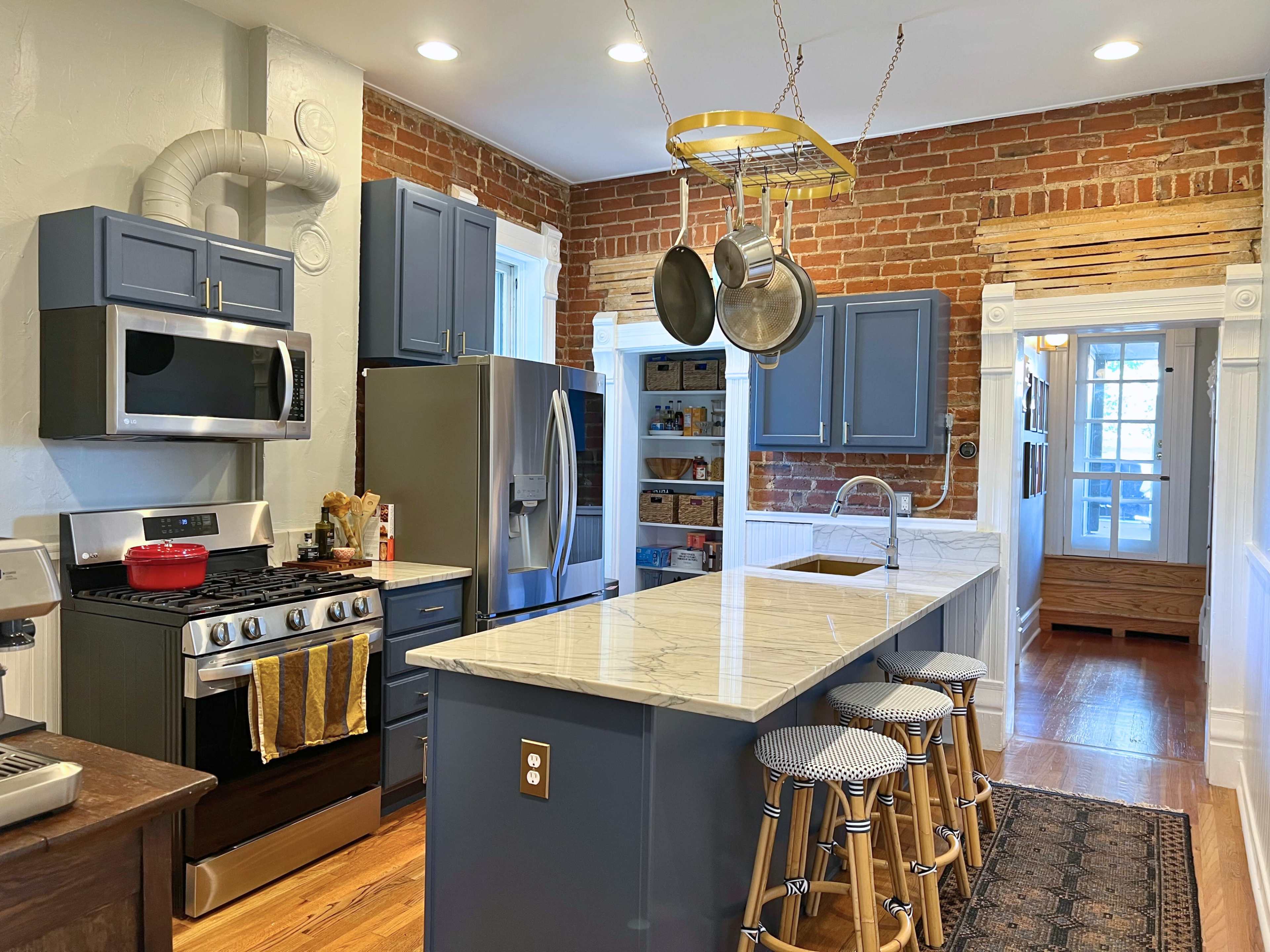 The image shows a modern kitchen featuring exposed brick walls, blue cabinetry, a large marble island, and stainless steel appliances.
