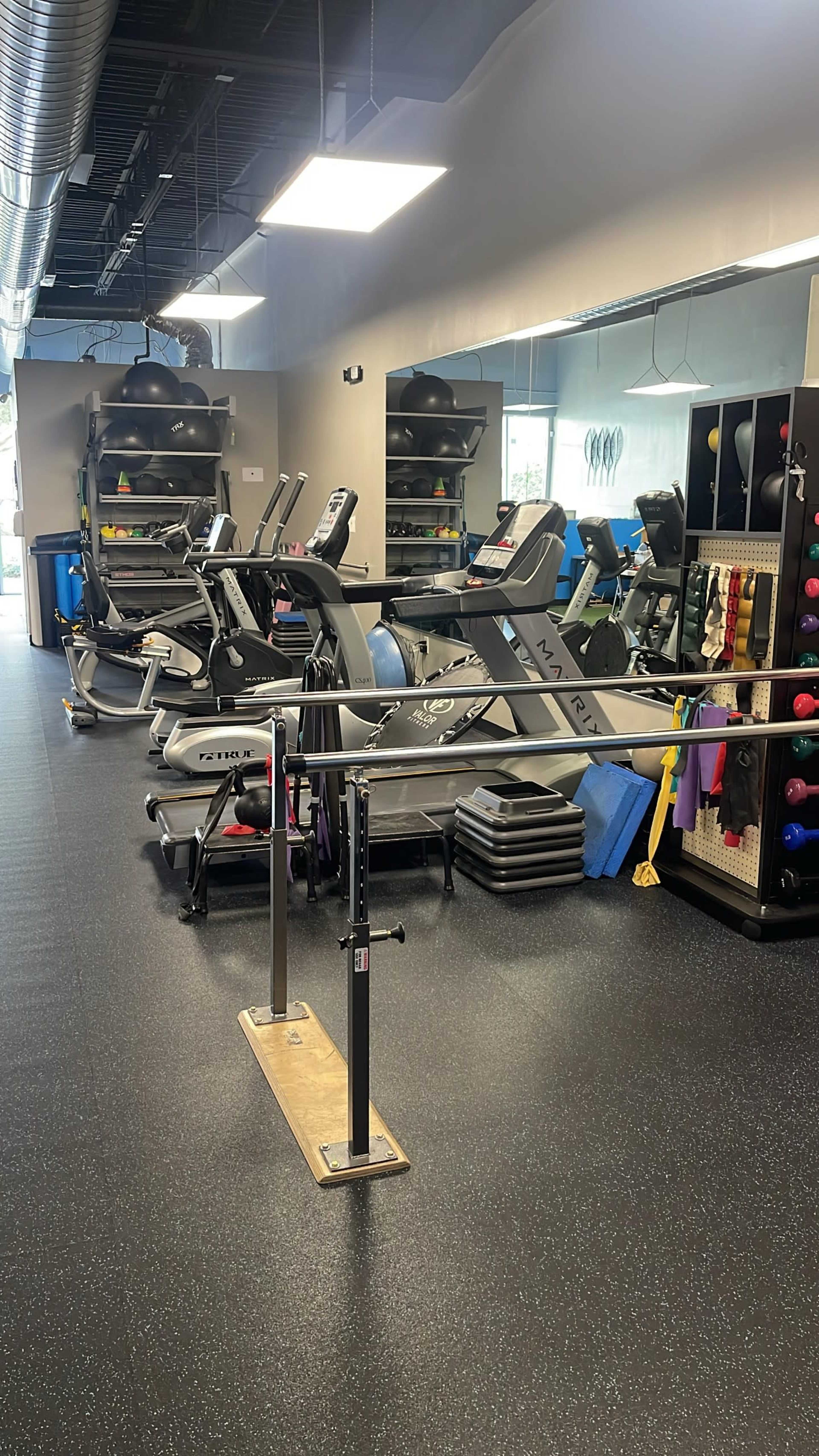 The image shows a gym with various exercise equipment, including treadmills, stationary bikes, and a balance bar, arranged on a rubberized floor.