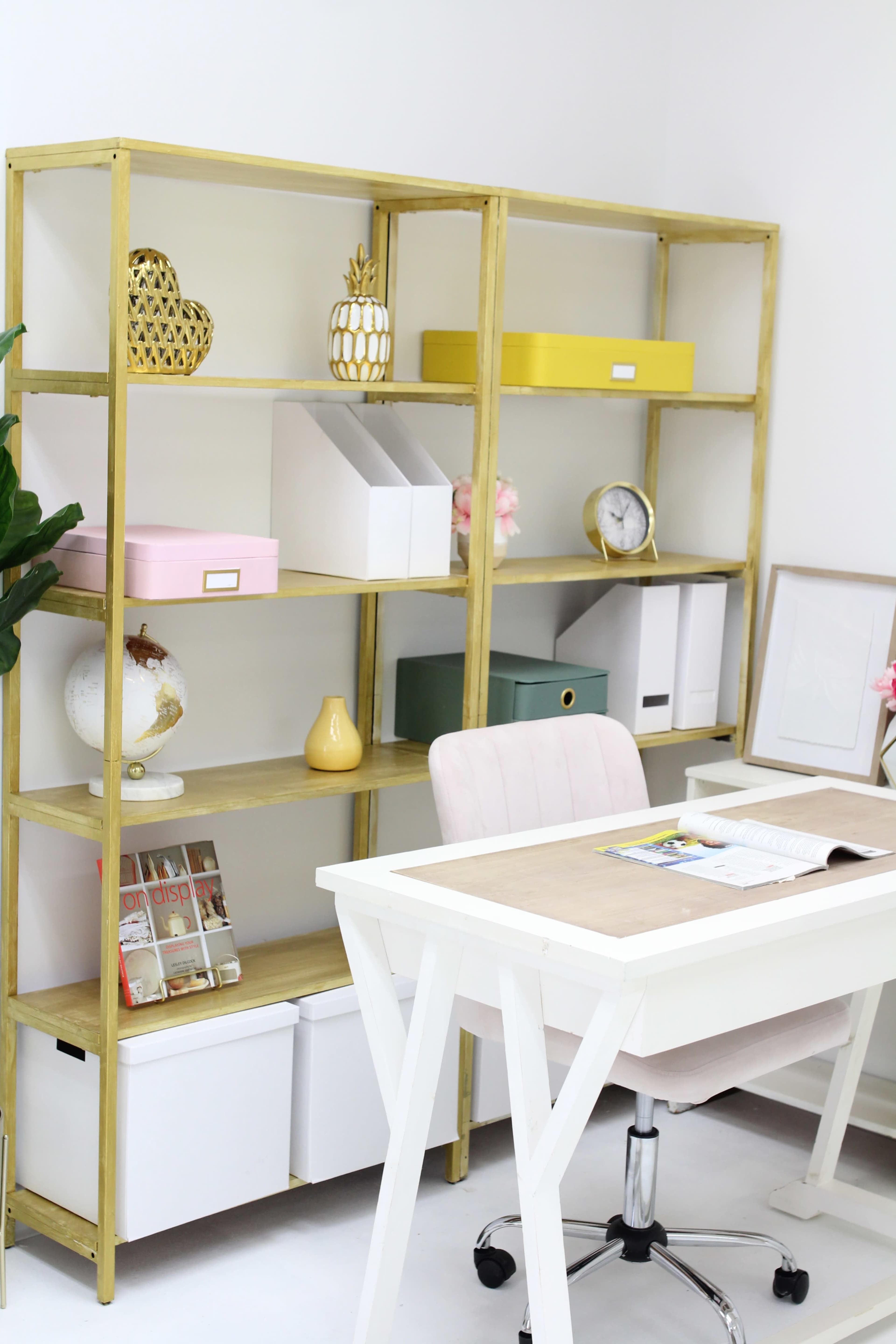A wooden bookshelf filled with decorative items and storage boxes stands next to a white desk and an upholstered chair in a bright office space.