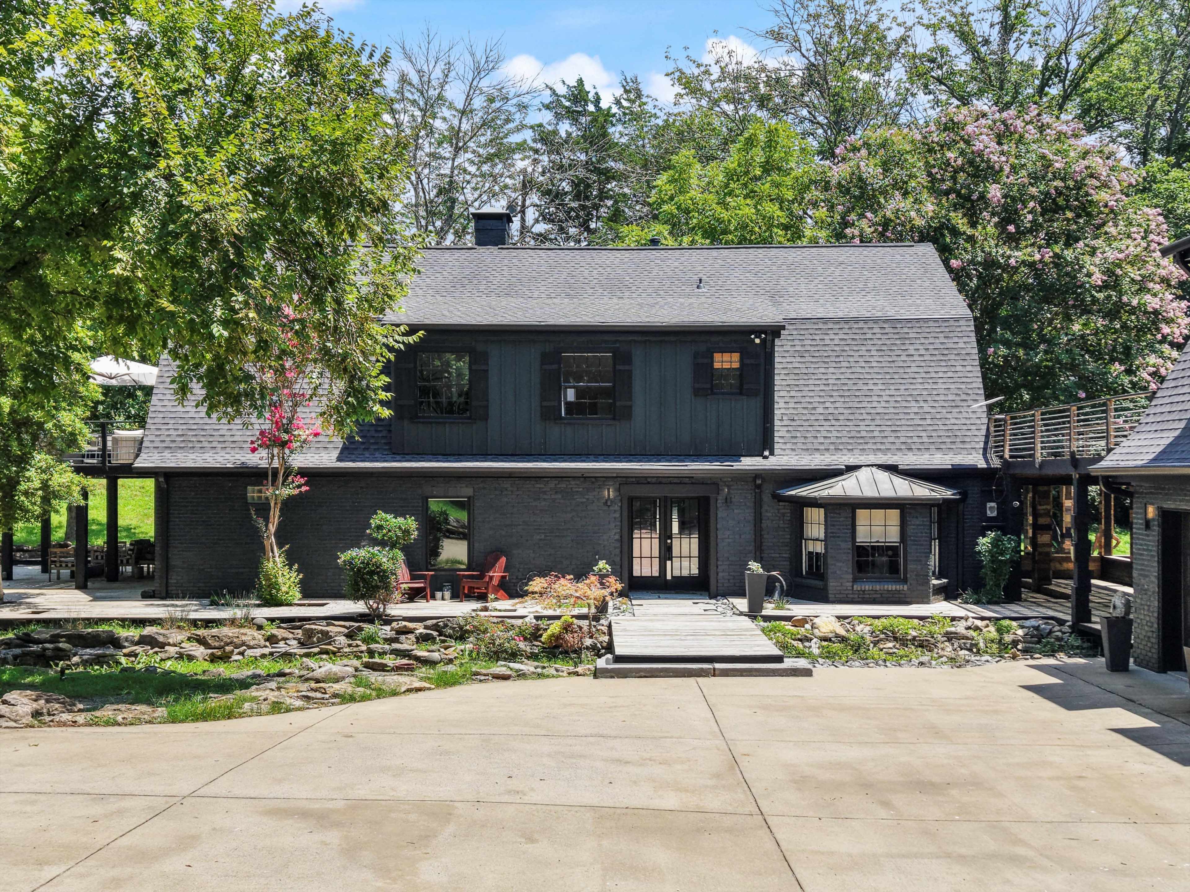 A modern black house with a sloped roof is surrounded by trees and features a stone pathway and outdoor seating area.