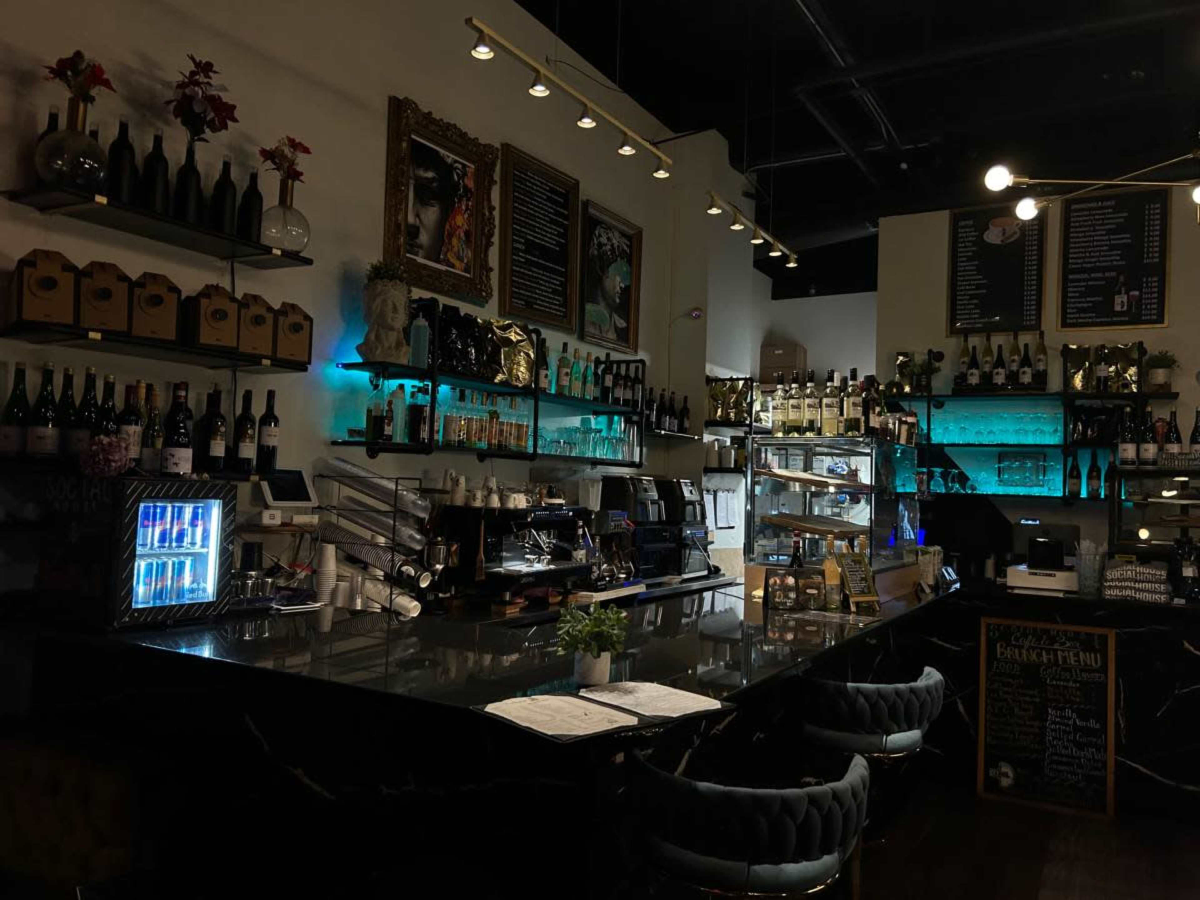 The image shows a dimly lit bar counter with shelves displaying various bottles and a coffee machine, accompanied by a menu board and decorative art on the walls.