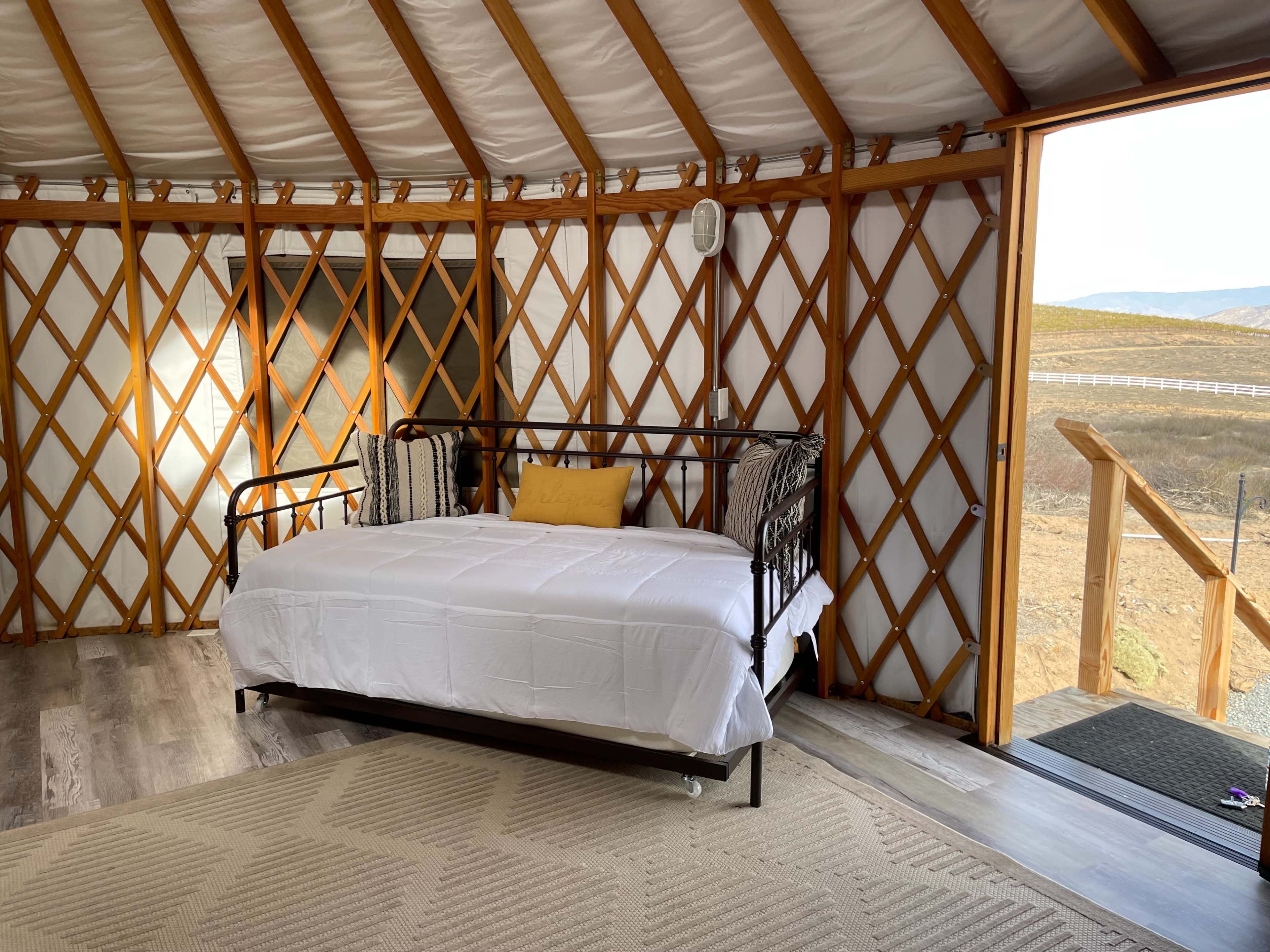 The interior of a yurt features a daybed with a white comforter, flanked by patterned pillows, and large windows showing a view of the landscape outside.