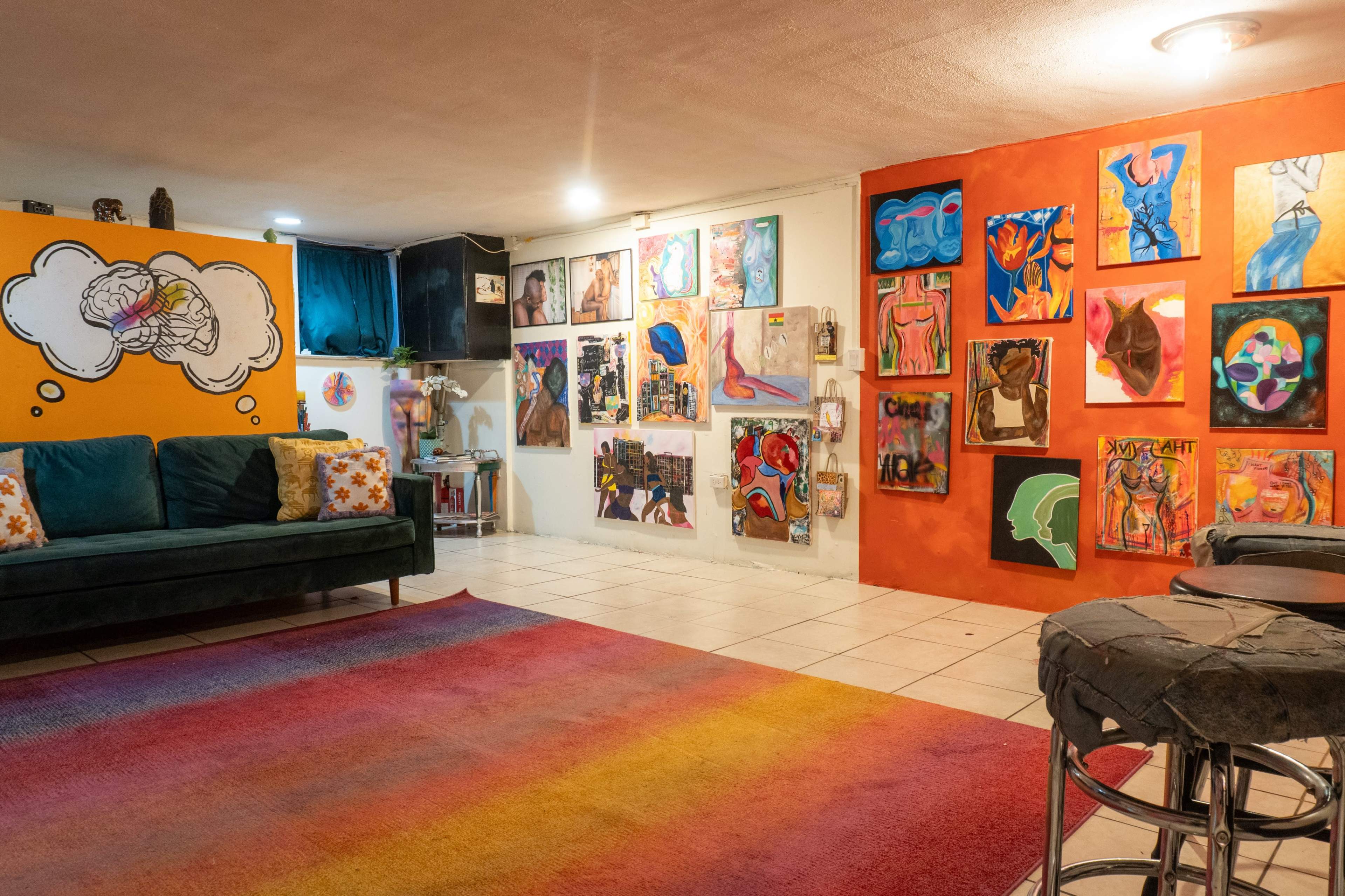 The image depicts a colorful art-filled room featuring a green couch, a vibrant orange wall with framed artwork, and a multi-colored rug on the tiled floor.