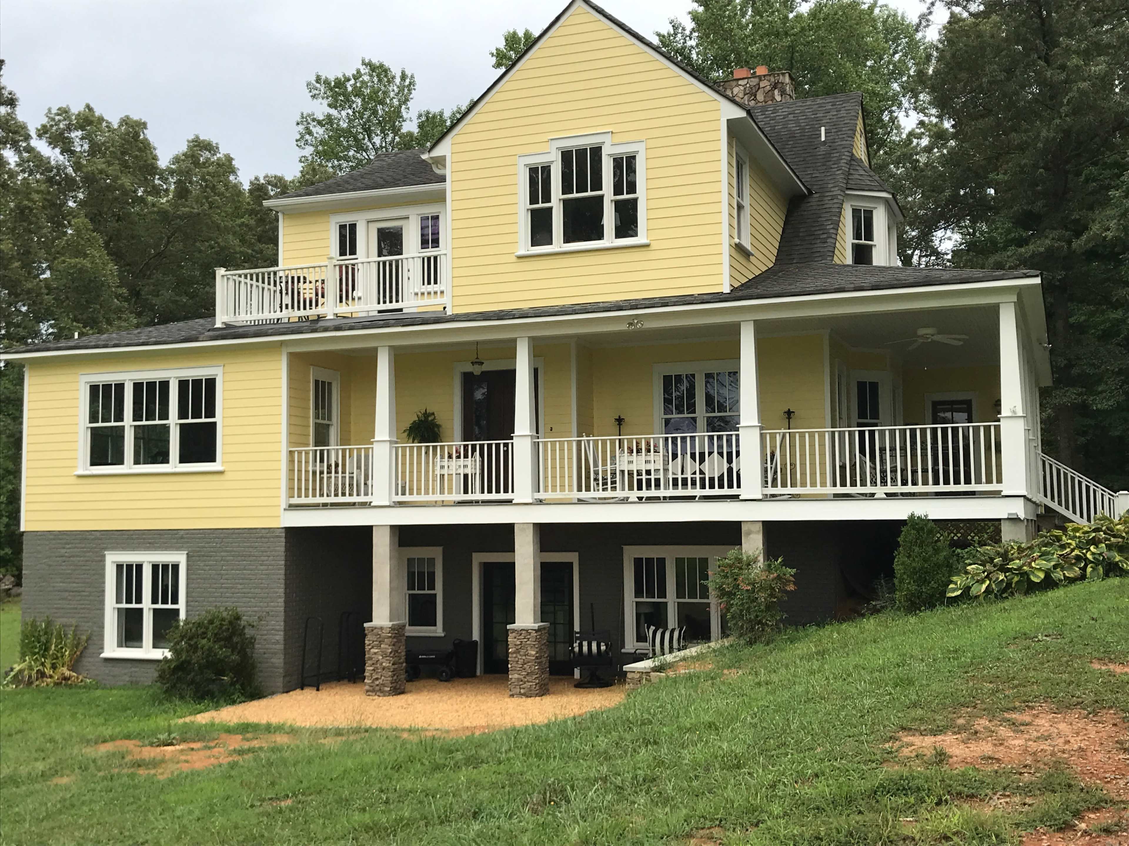 A two-story yellow house with a grey stone foundation and a front porch is situated on a grassy slope surrounded by trees.