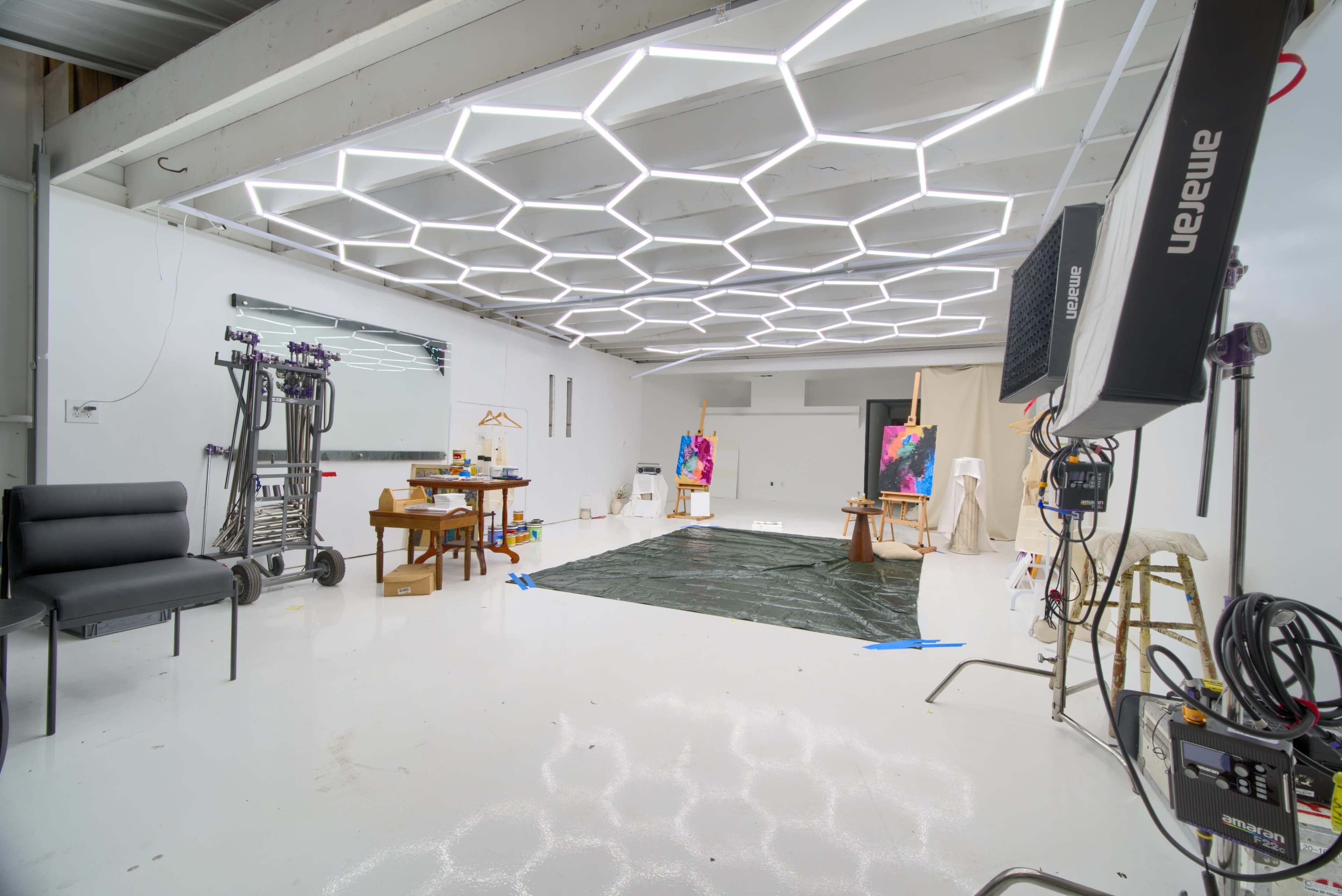 The image shows a bright, spacious studio with geometric ceiling lights, a black tarp on the floor, and various art supplies and equipment placed around the room.