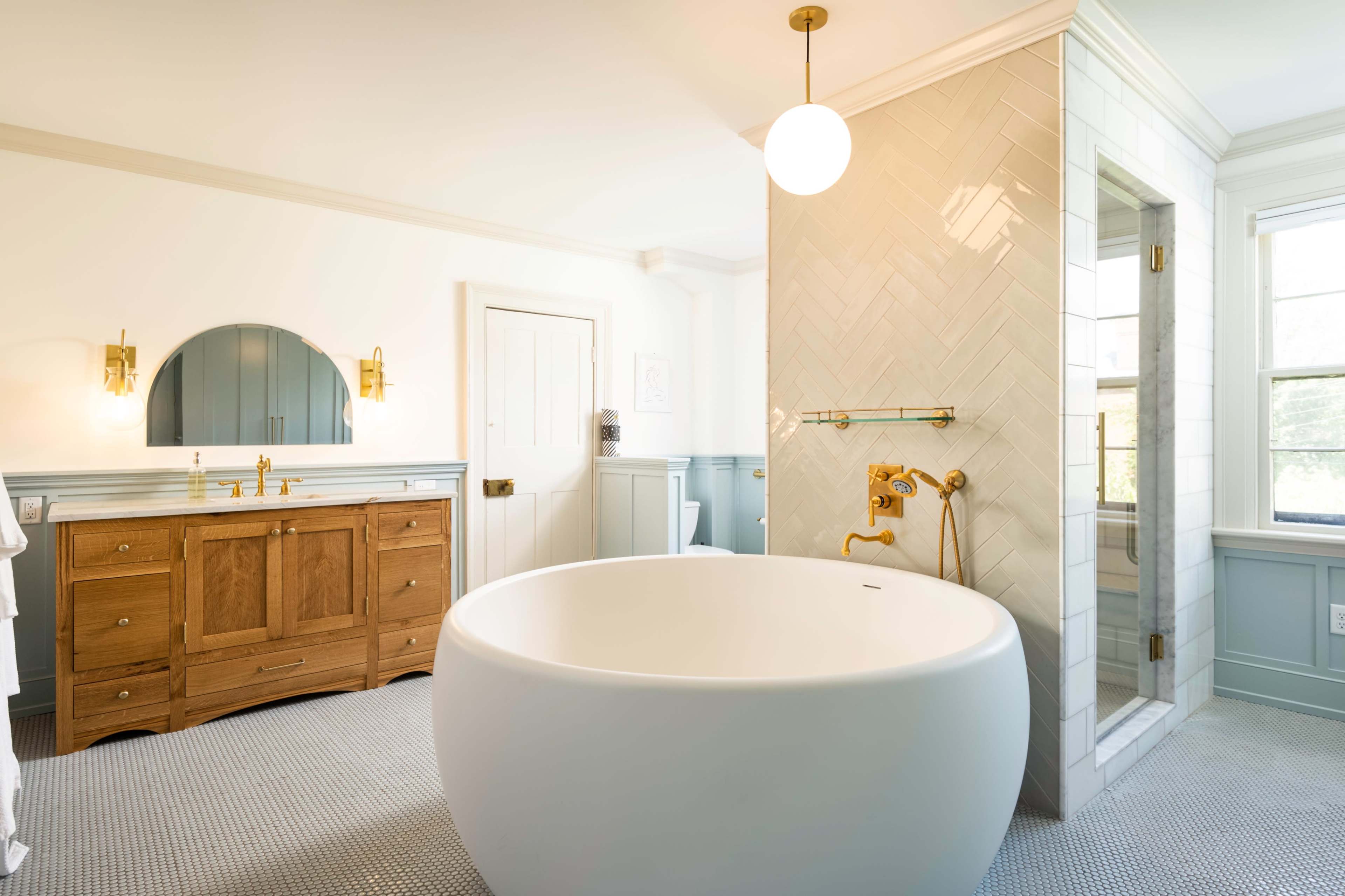 The bathroom features a freestanding white bathtub, a wooden vanity with gold fixtures, and a glass shower enclosed with herringbone tile.