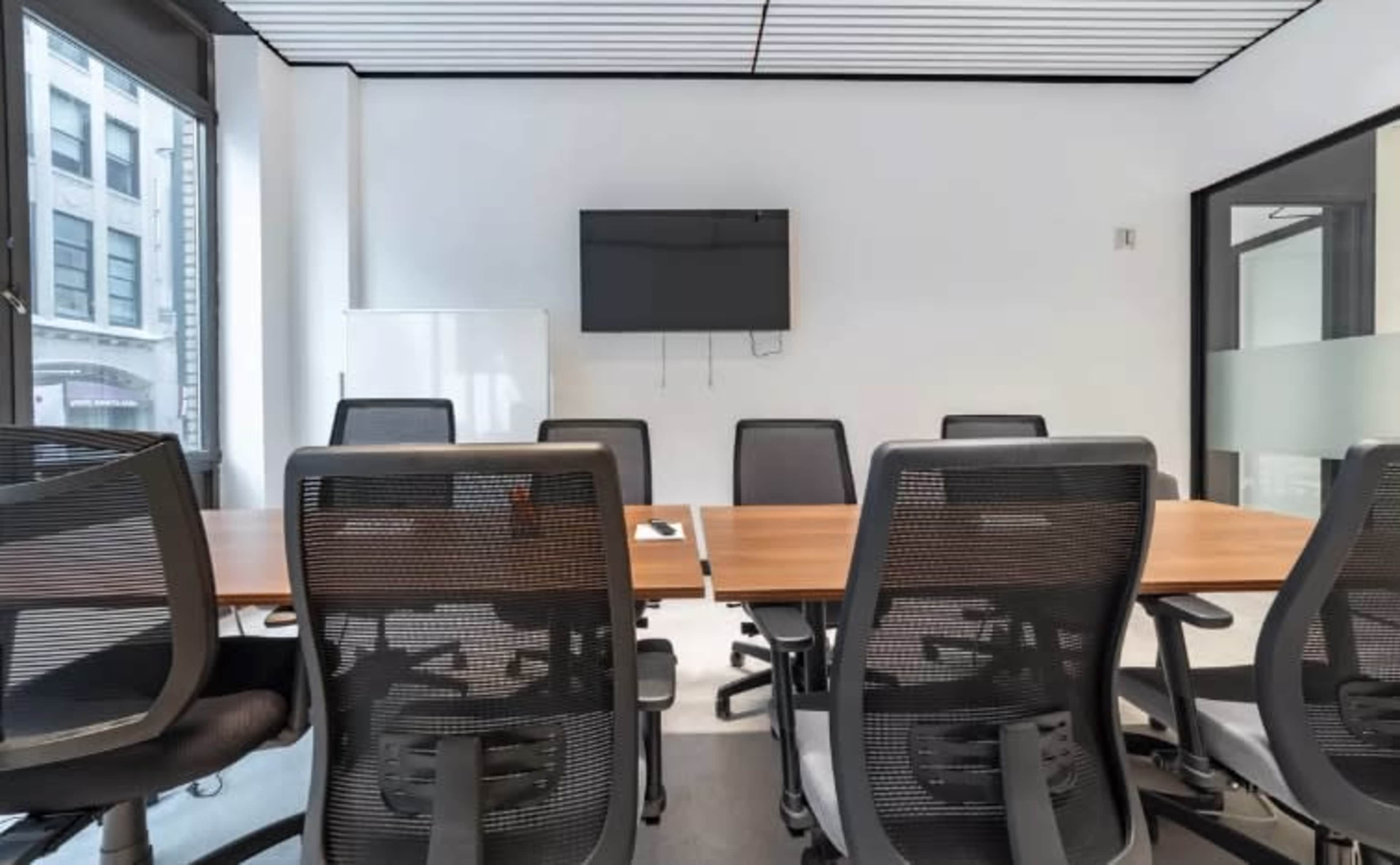A modern conference room features a long table surrounded by ergonomic chairs, with a large screen mounted on the wall.