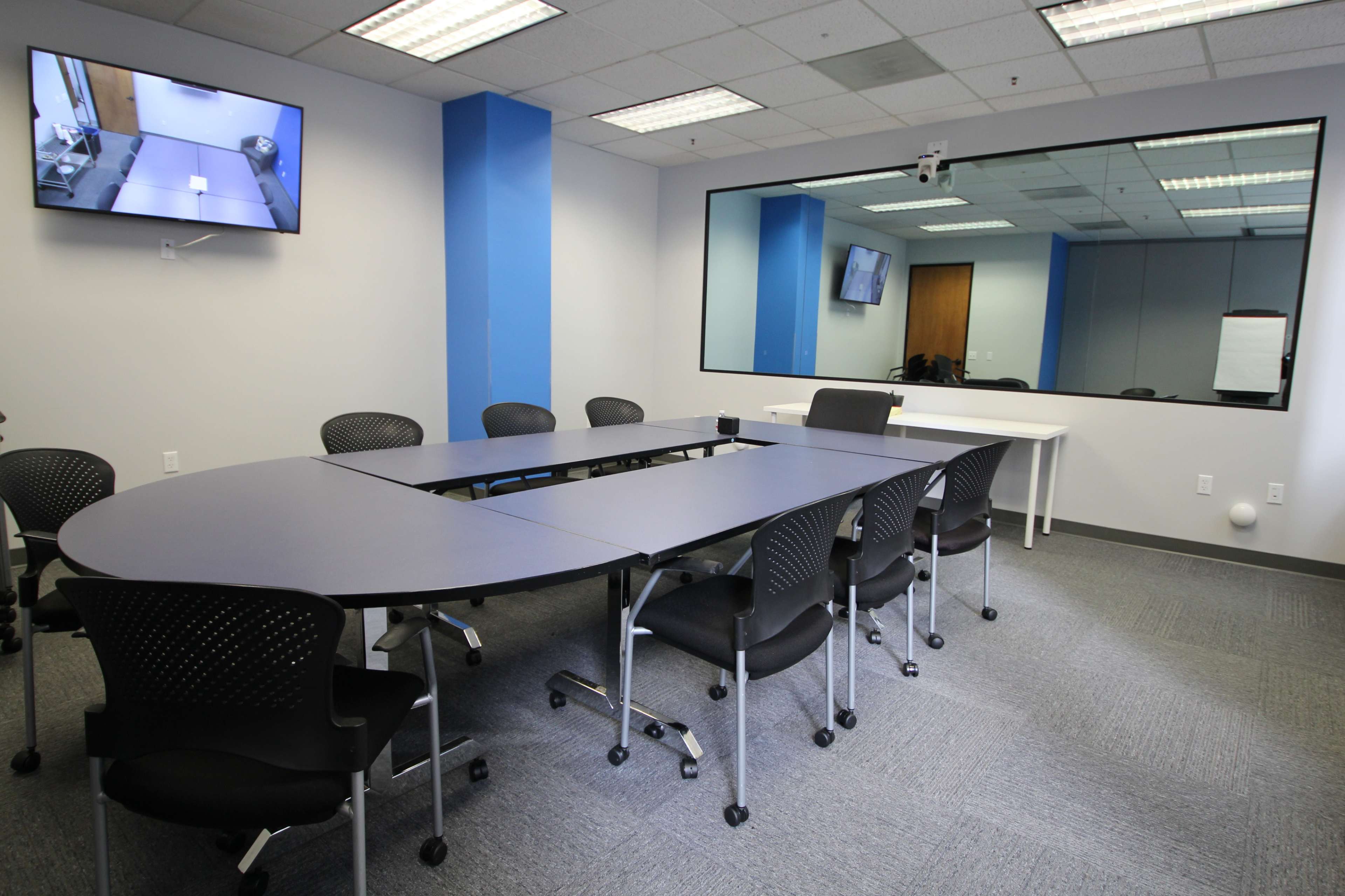 A conference room with an oval table surrounded by black chairs, a large mirror, a television mounted on the wall, and a white flip chart holder.