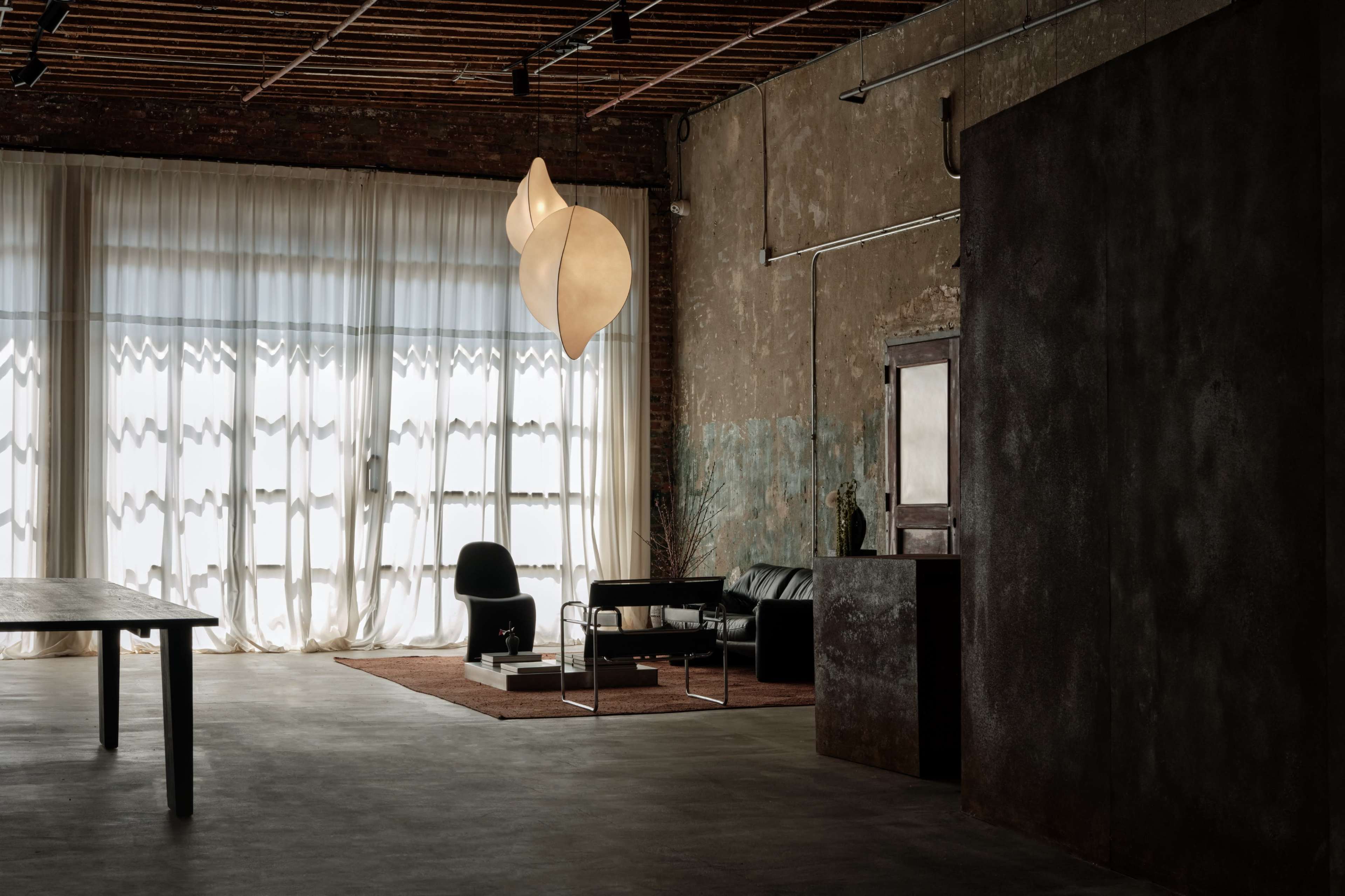 The image shows a modern industrial interior space featuring a black leather sofa, a contemporary light fixture, and large windows draped with white curtains.