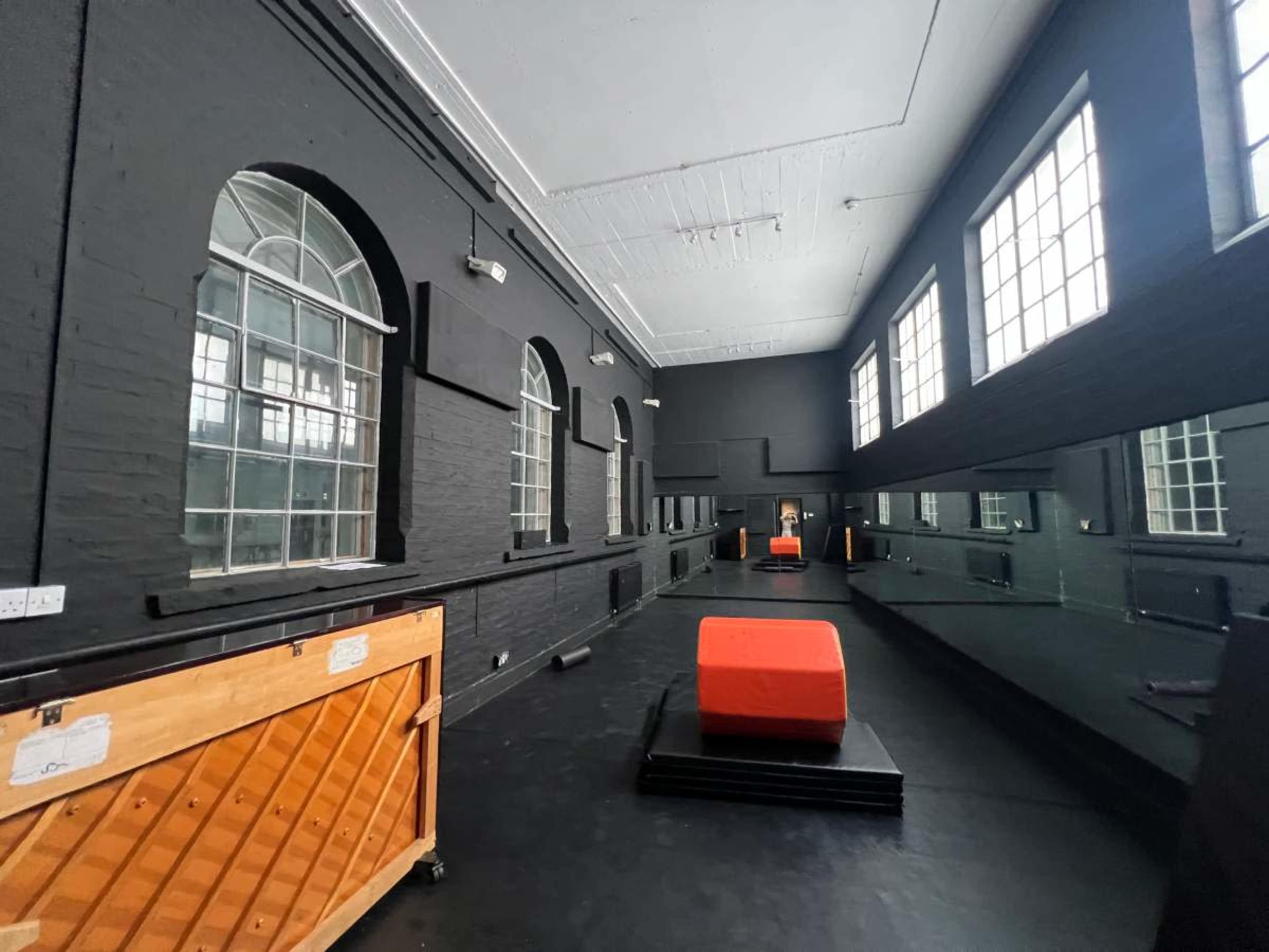 The image shows a spacious dance studio with black walls, large windows, and a mirrored wall, featuring an orange block in the center.