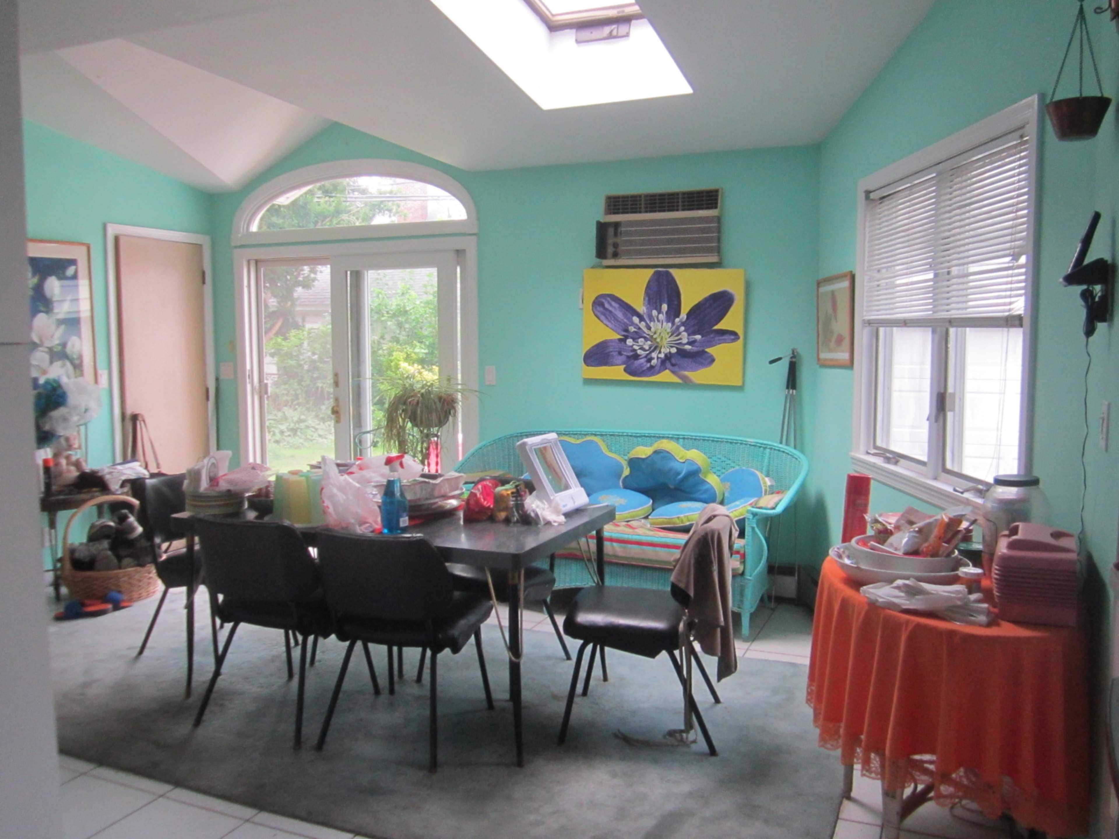A bright room features a turquoise wall, a large skylight, a dining area with chairs, a table cluttered with items, and a colorful piece of floral art.
