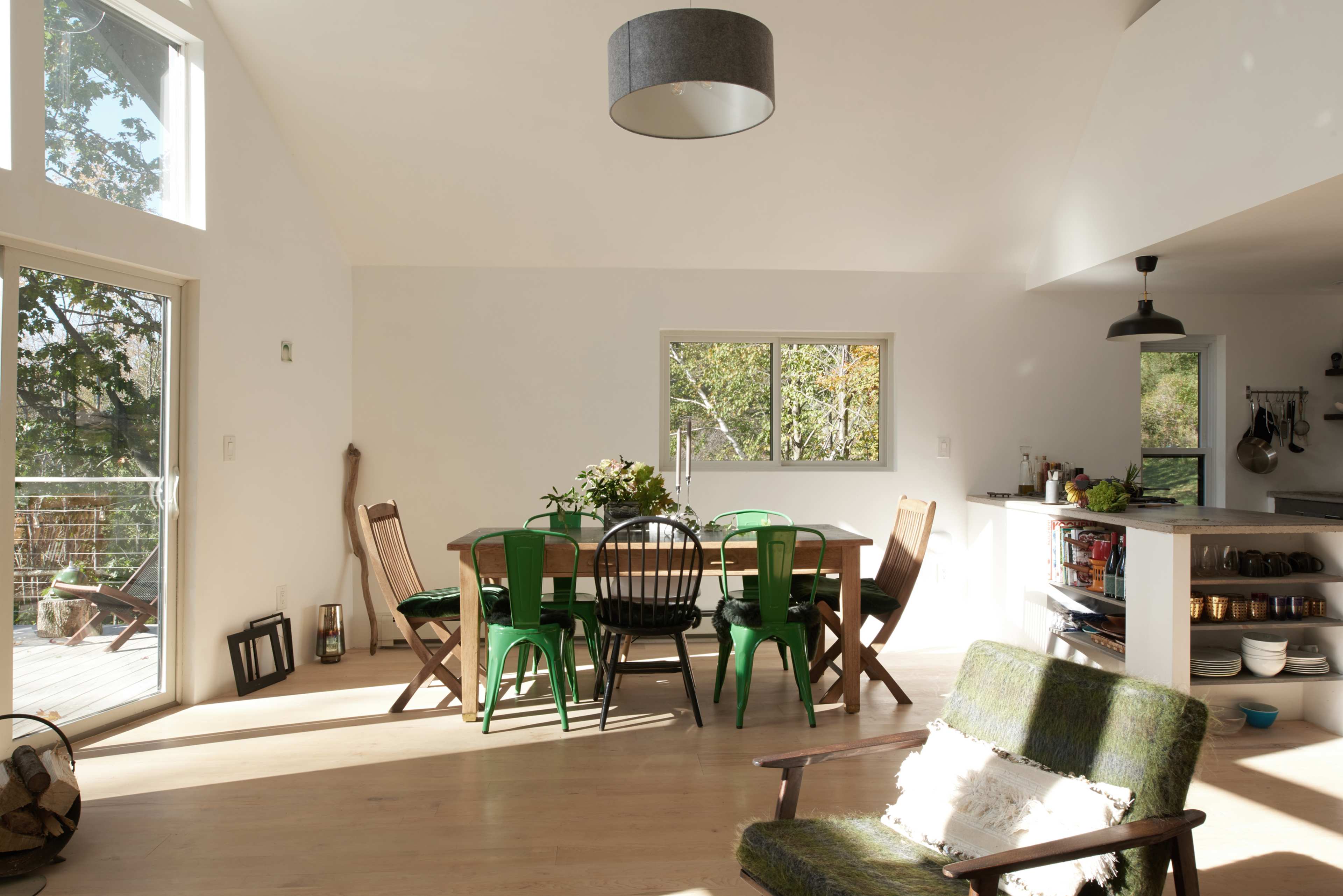 A light-filled dining area features a wooden table surrounded by chairs and a green upholstered armchair, with a view of trees through large windows.