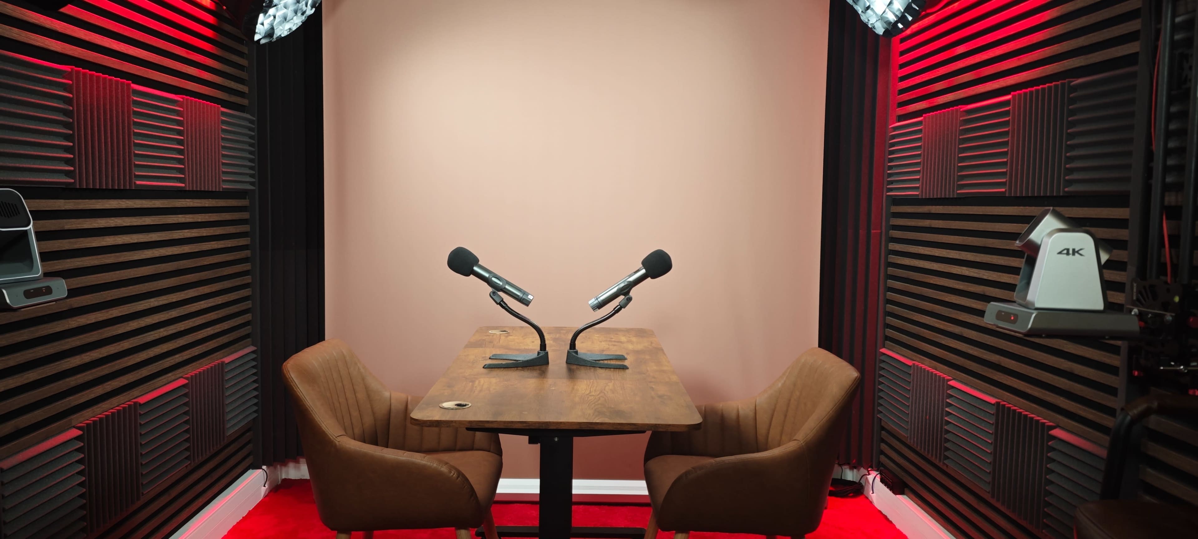 A podcast studio features a wooden table with two microphones and two brown chairs against a pink wall, accented by soundproofing panels and red lighting.