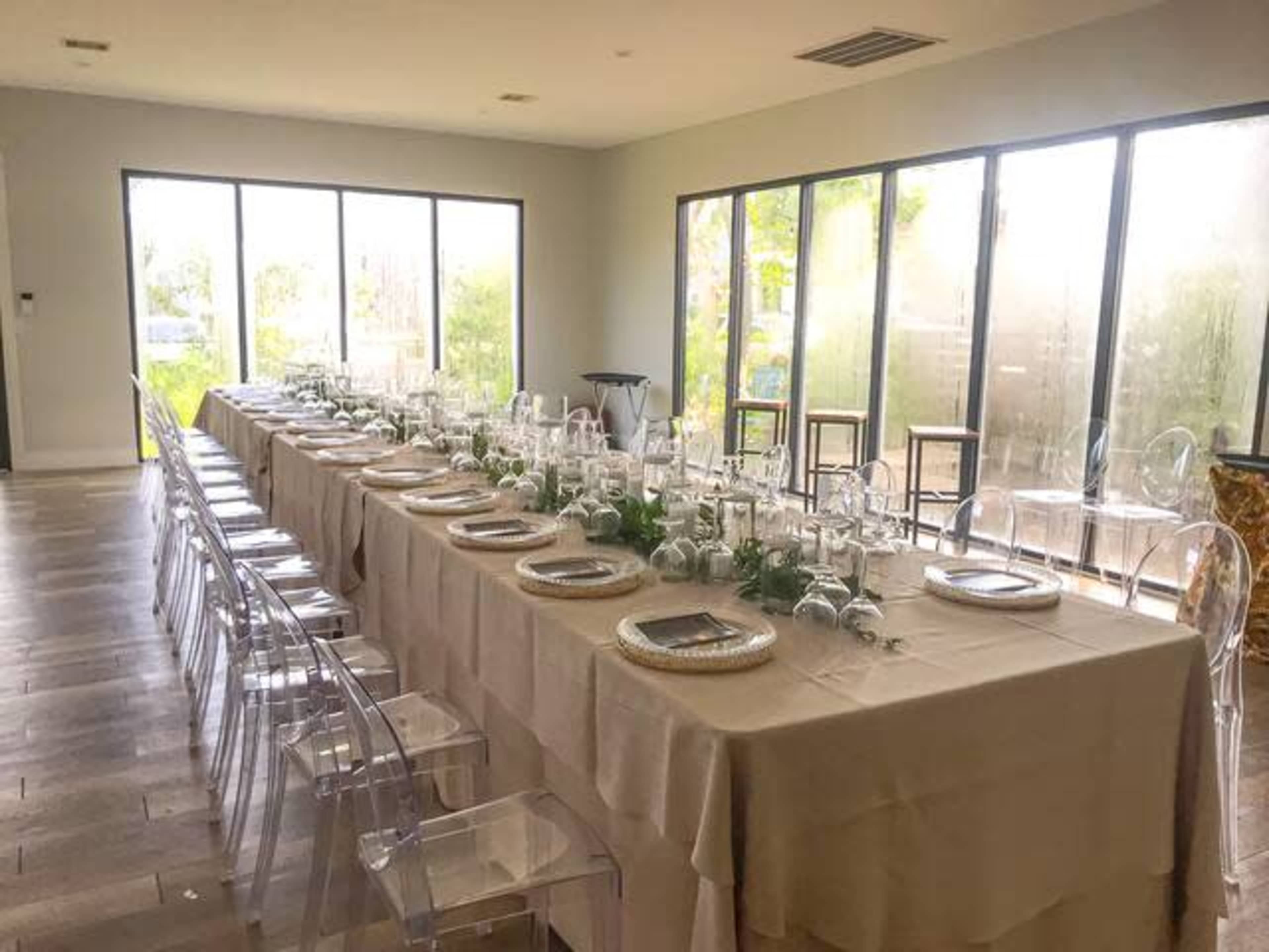 A long dining table is set with glassware and tableware, surrounded by clear acrylic chairs, in a brightly lit room with large windows.