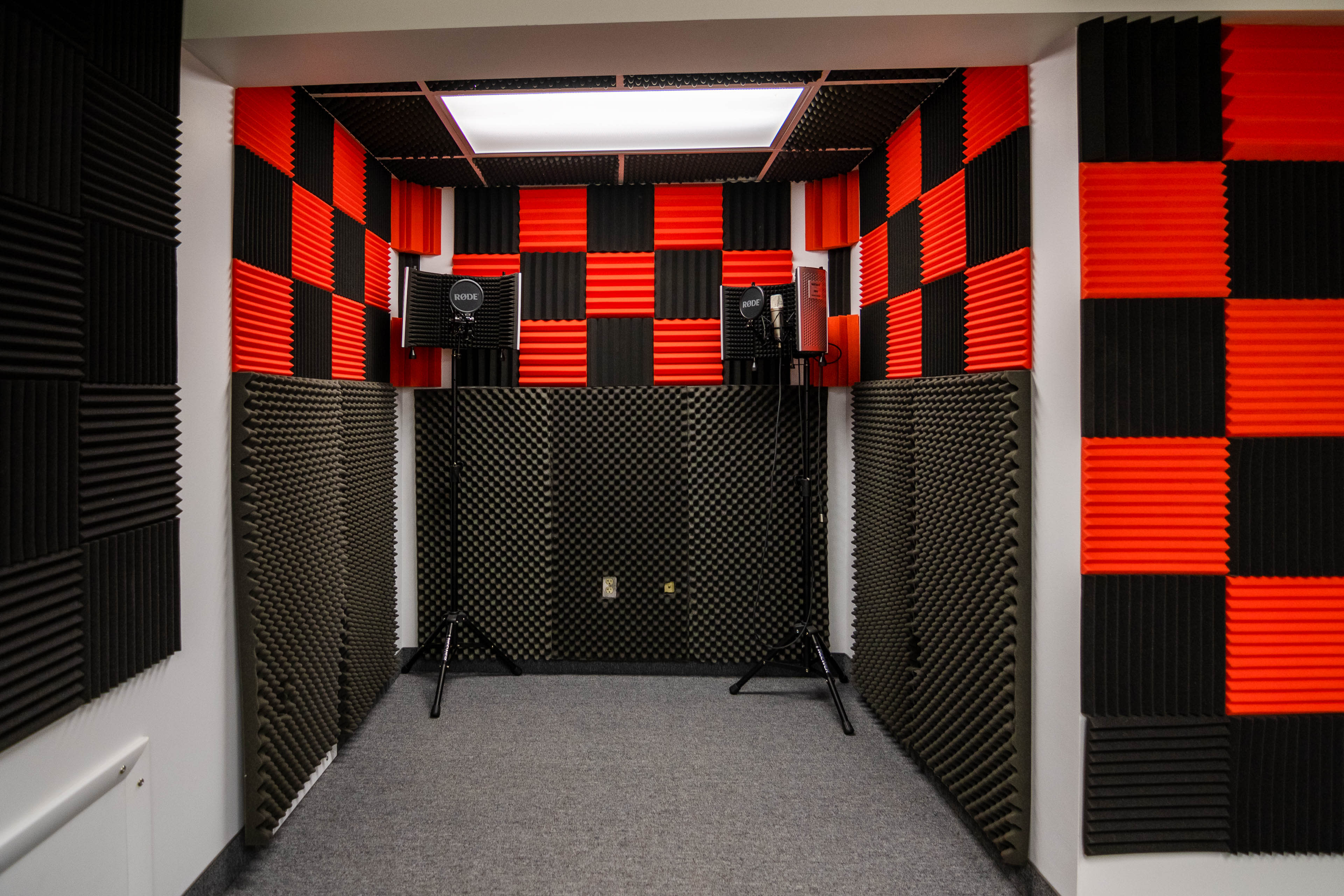 The image shows a soundproof recording studio featuring black and red acoustic foam tiles on the walls and two microphone stands positioned in the corners.